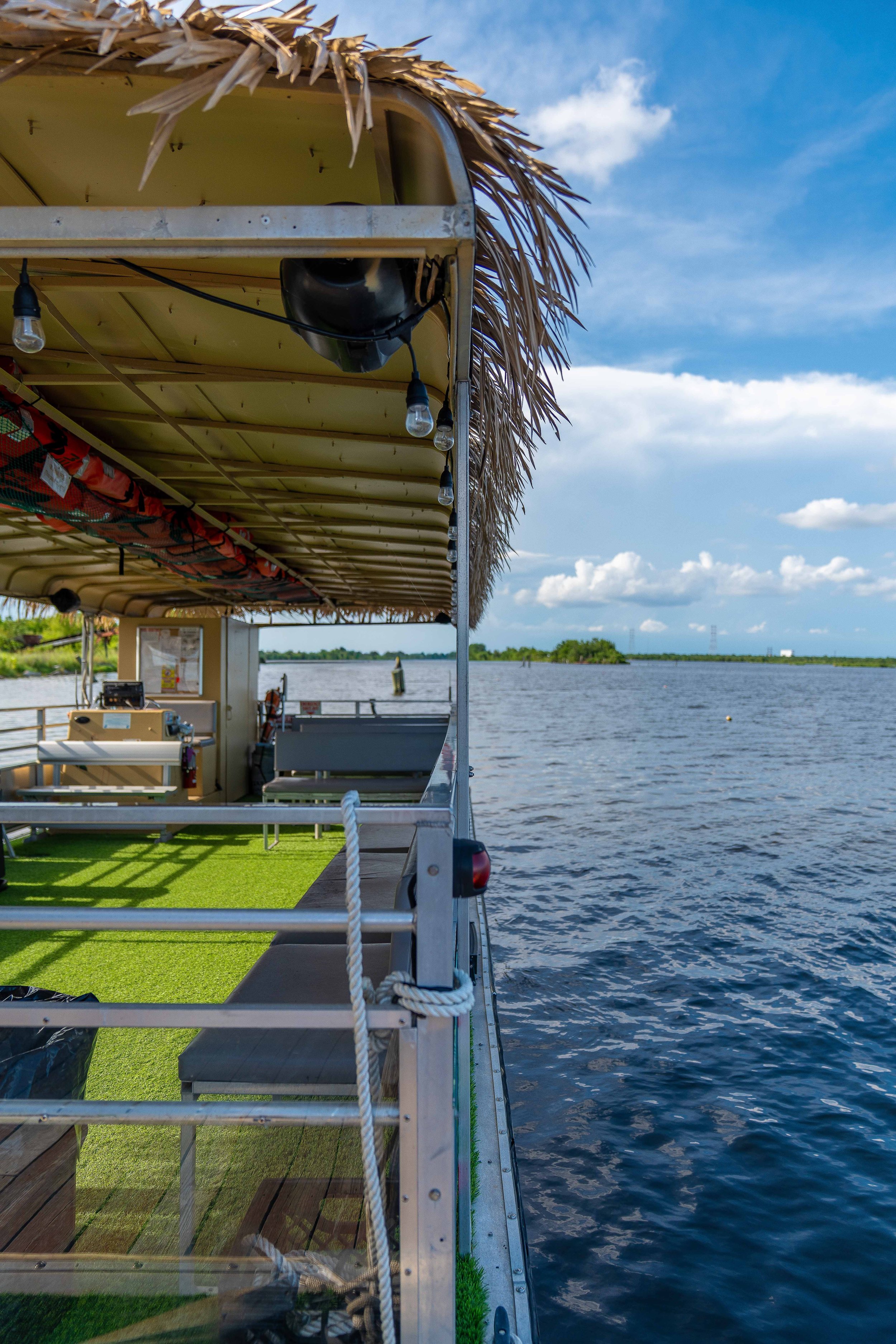 A boat with a thatched roof and string lights is floating on a river, with a grassy area inside the boat, and a clear sky with a few clouds overhead.