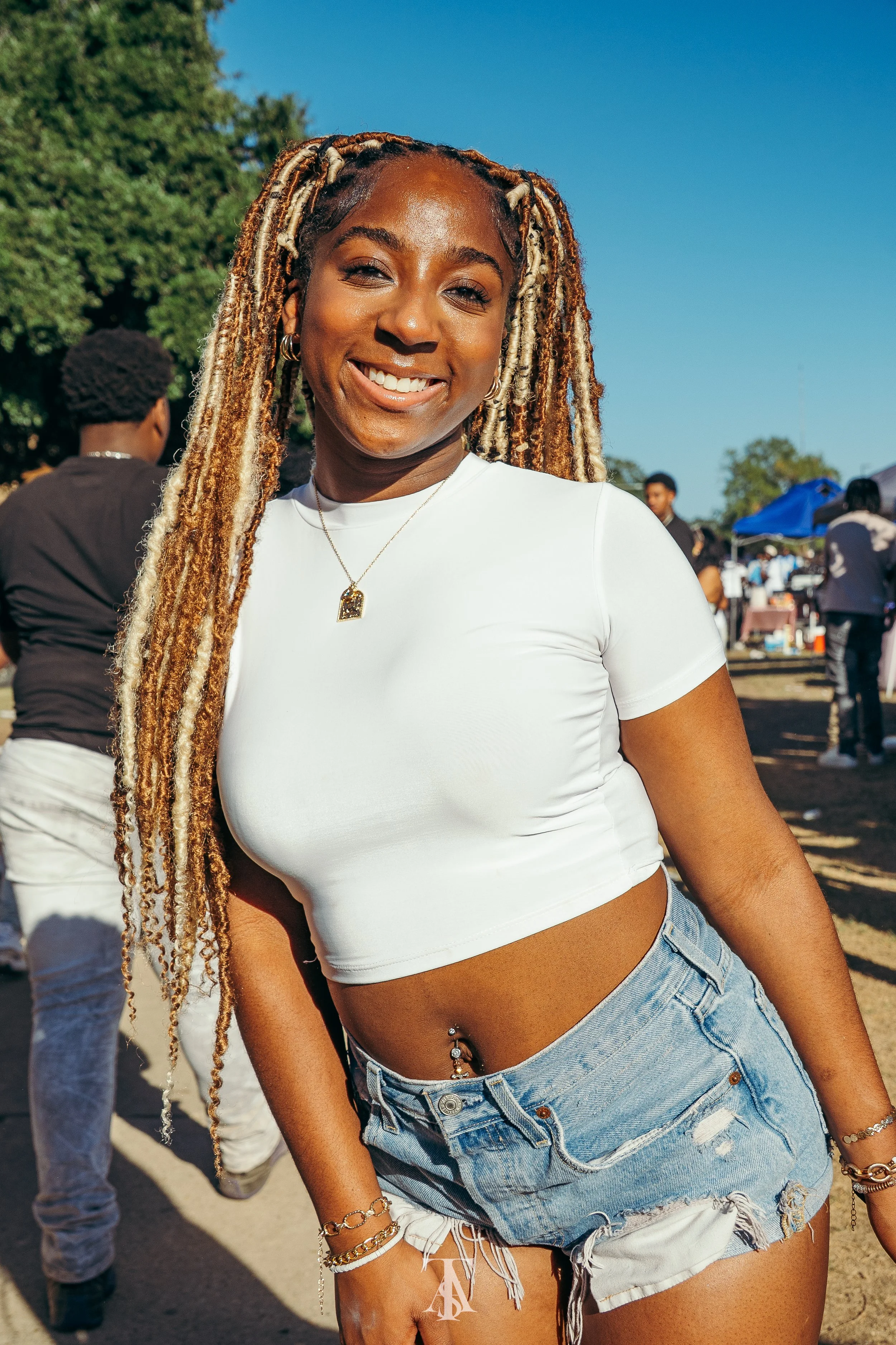 Smiling young woman with long blonde dreadlocks, wearing a white cropped t-shirt and distressed denim shorts, at an outdoor event on a sunny day.