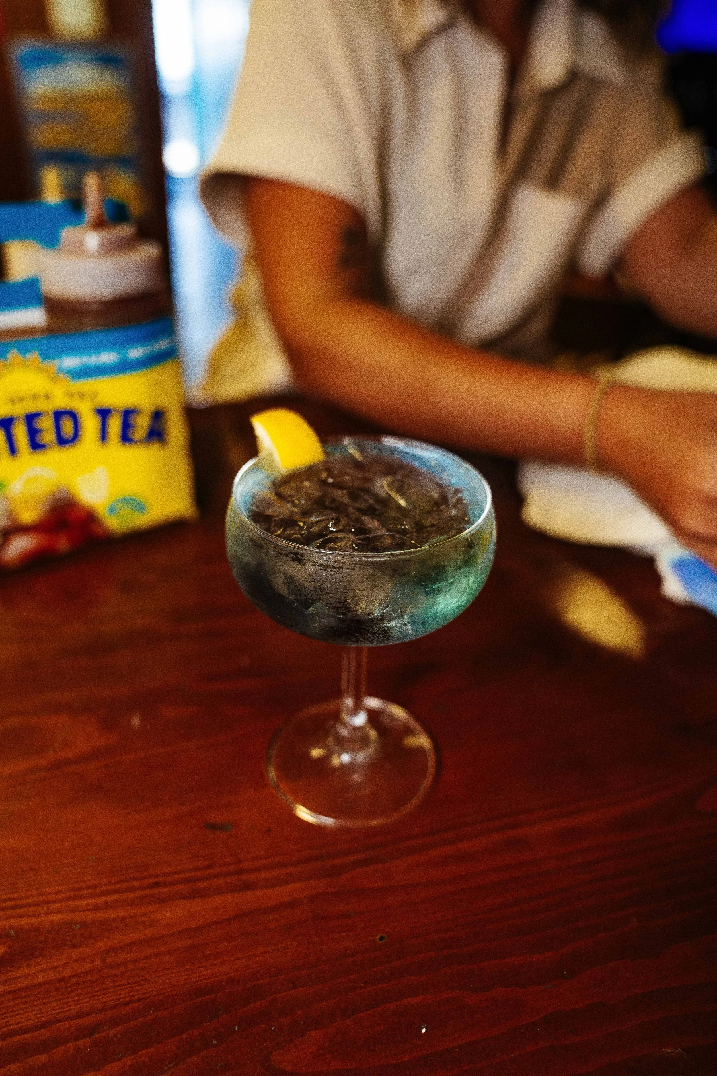 A glass of dark soda with ice and lemon on a wooden table. In the background, a person is sitting at the table, partially visible, with a colorful bag of iced tea and some other items nearby.