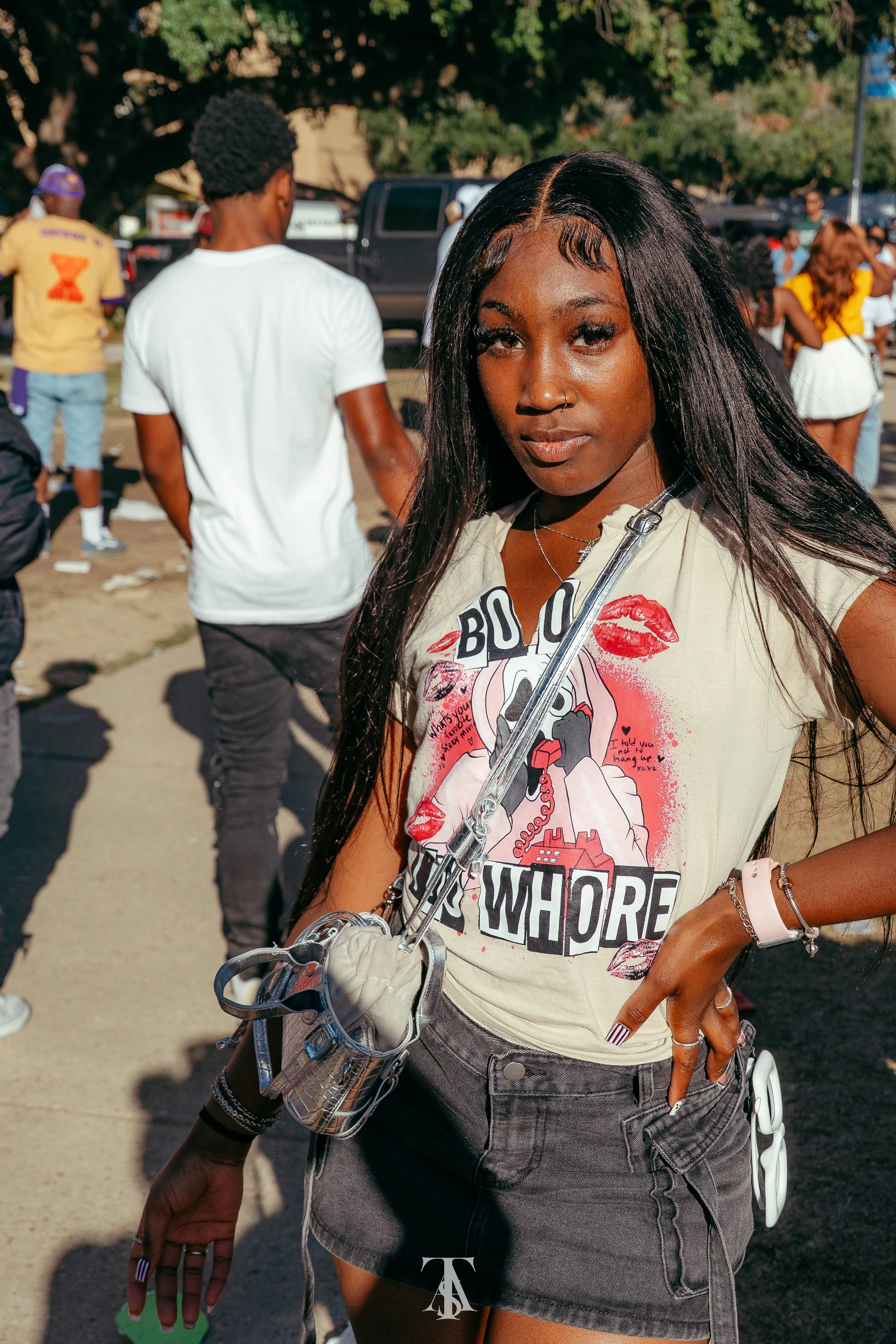 A young woman with long black hair, wearing a graphic t-shirt and a black denim skirt, stands outdoors at what appears to be a social event with people in the background.