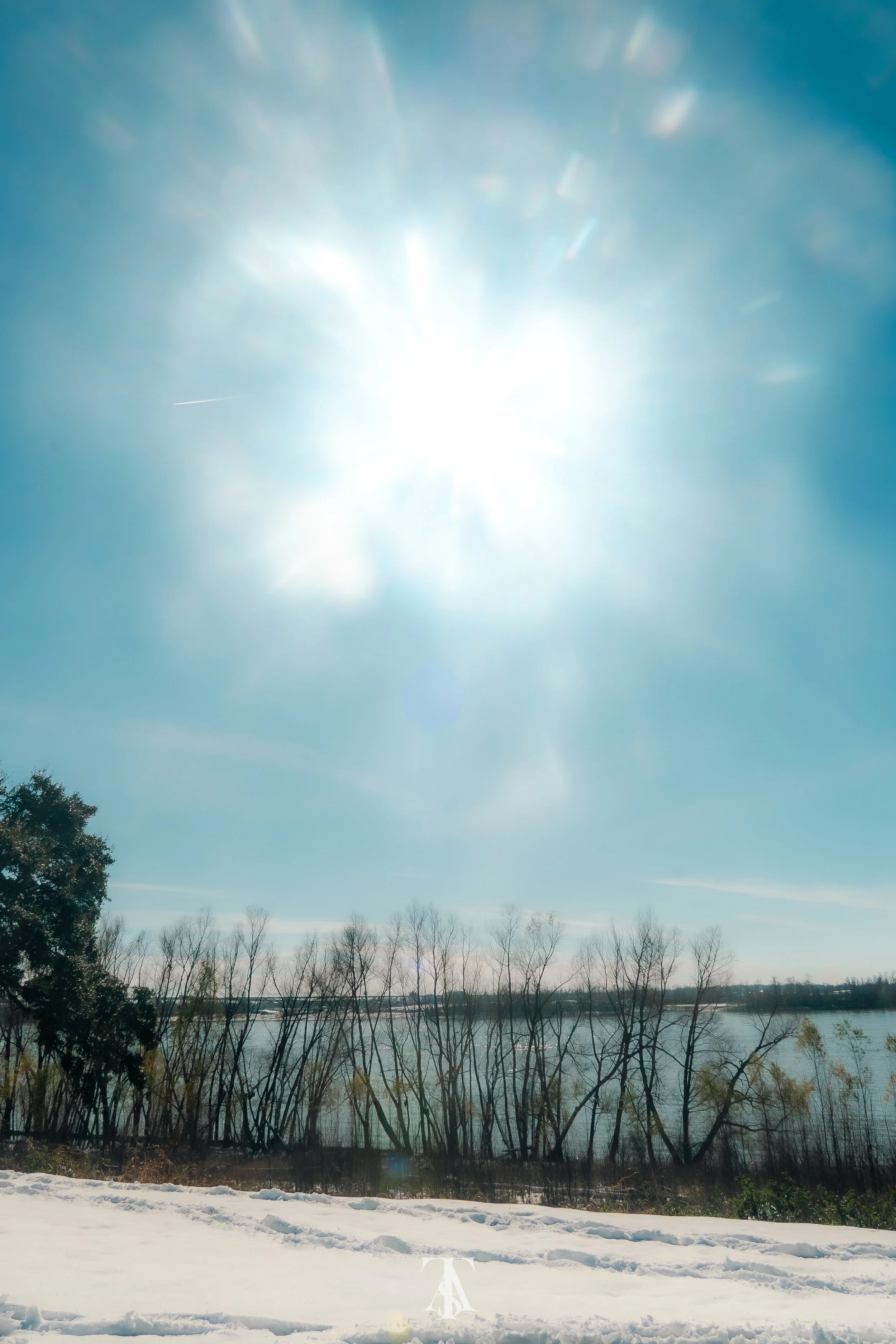 Bright sun shining in a clear blue sky above leafless trees near a body of water, with snow on the ground.