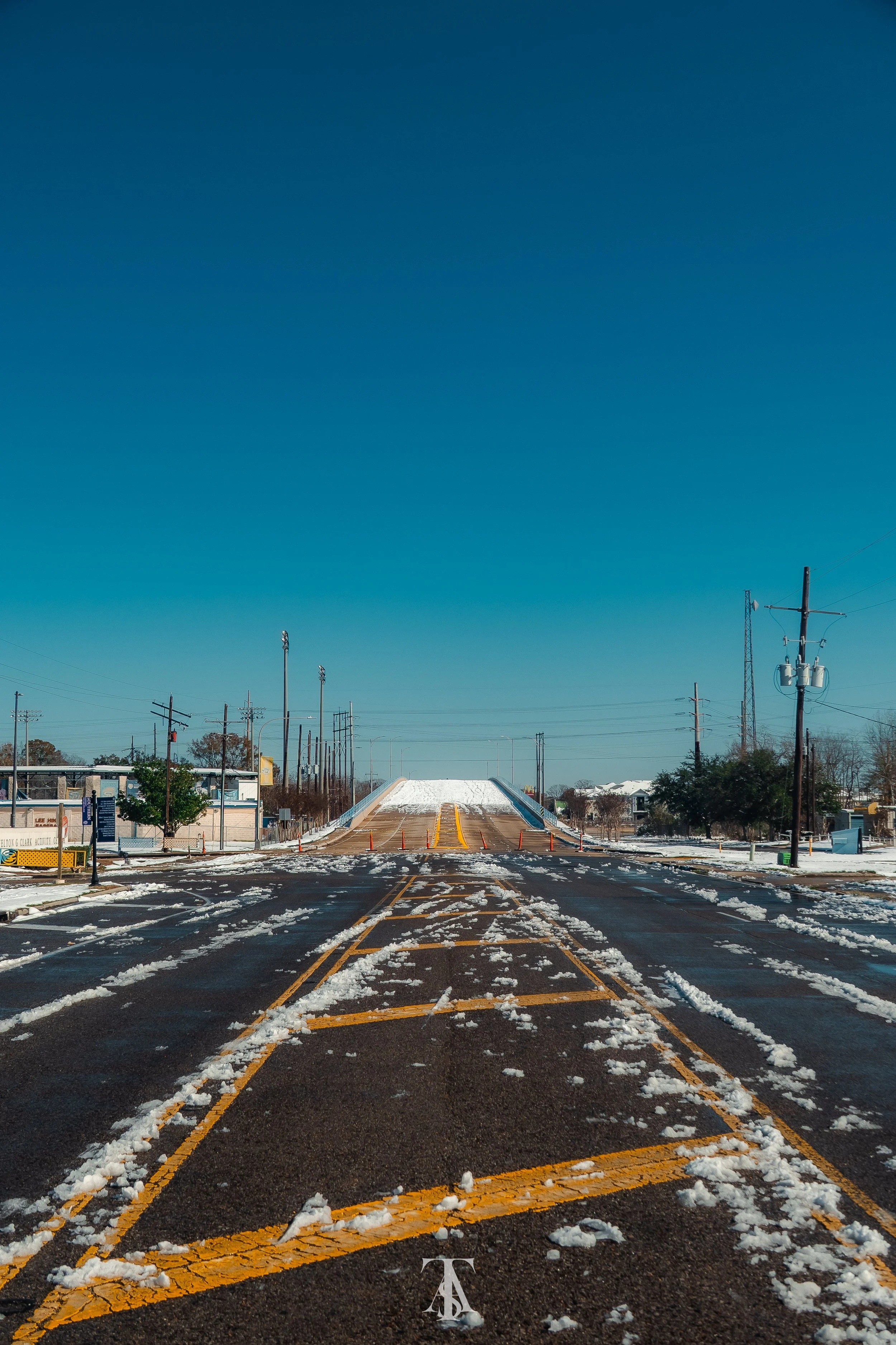 An empty street with snow and slush, leading to a bridge or ramp in the background, with utility poles on the sides and a clear blue sky.