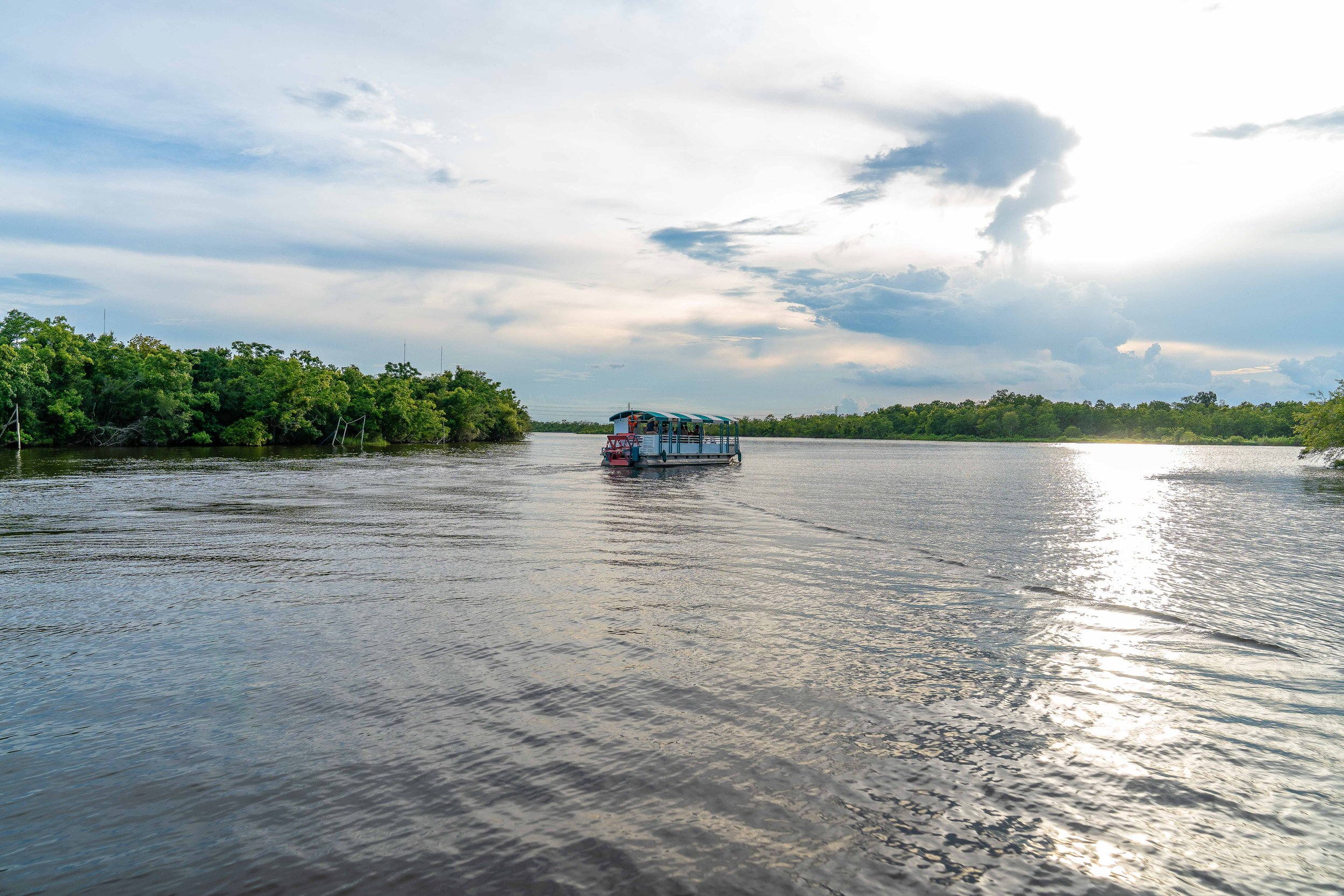 A boat floating on a river with trees on the riverbank under a partly cloudy sky.