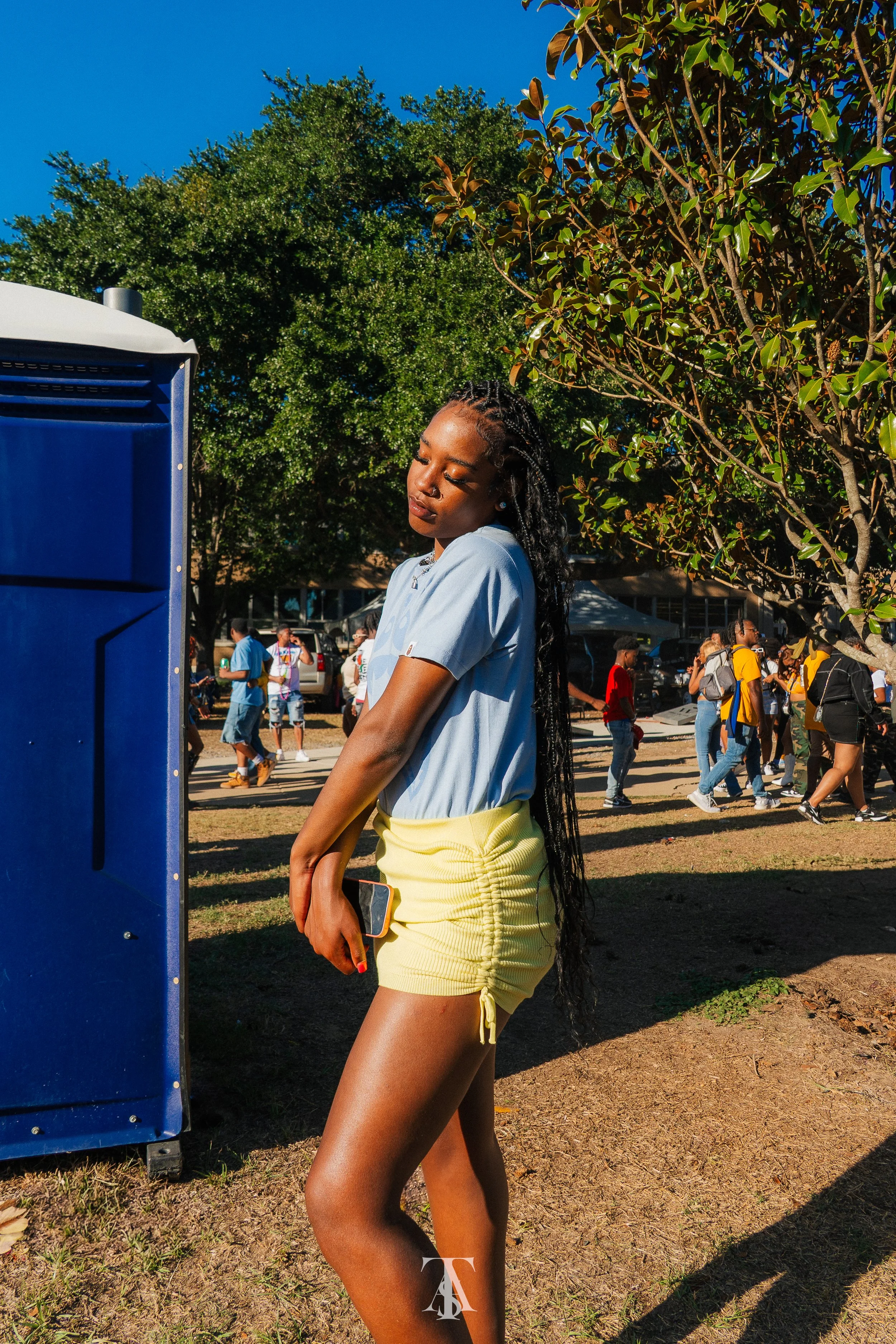 A young woman with long braided hair in a light blue t-shirt and yellow ruched shorts stands outdoors, holding a phone in her right hand, with her eyes closed, near a blue vending machine, with a group of people in the background at a park or festiva