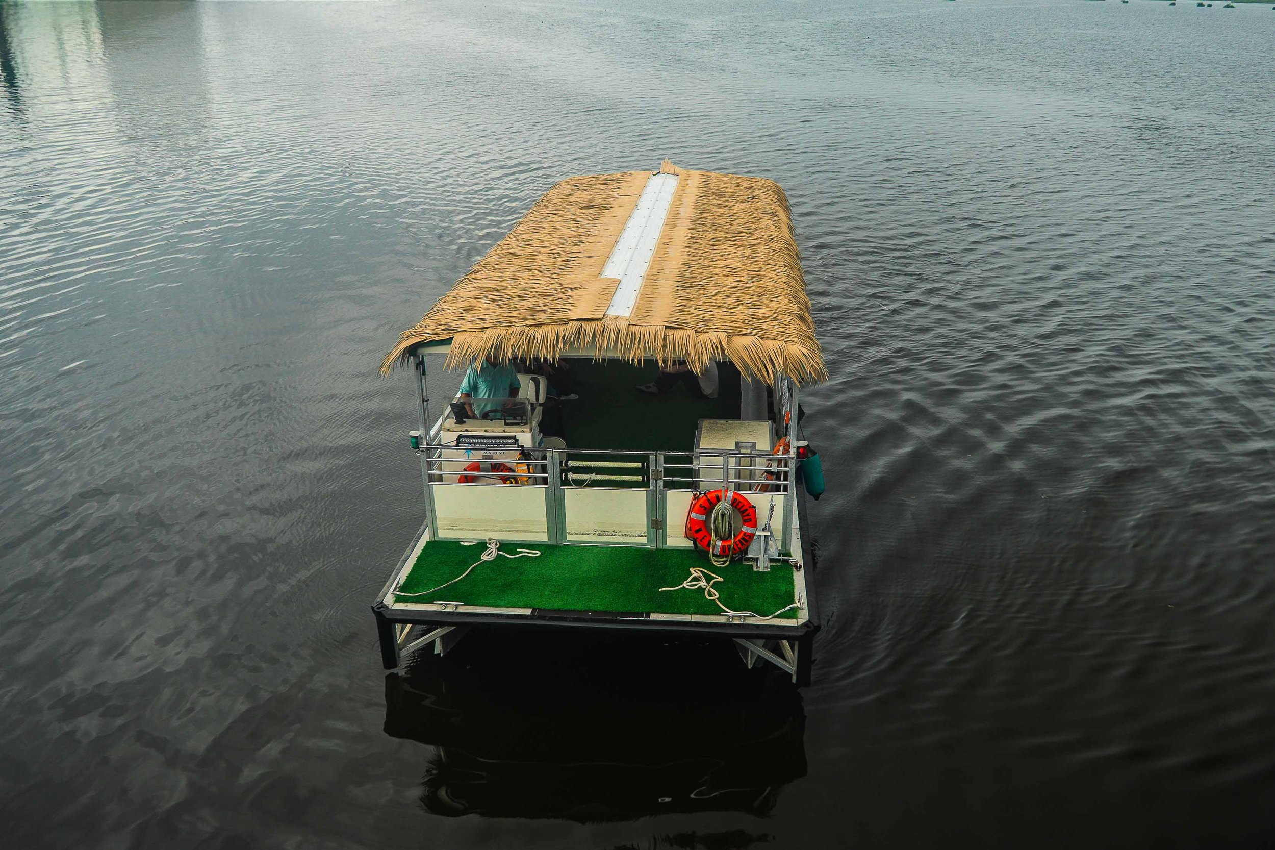 A boat with a straw roof and green carpet floating on calm water, with a person in a teal shirt visible at the front.