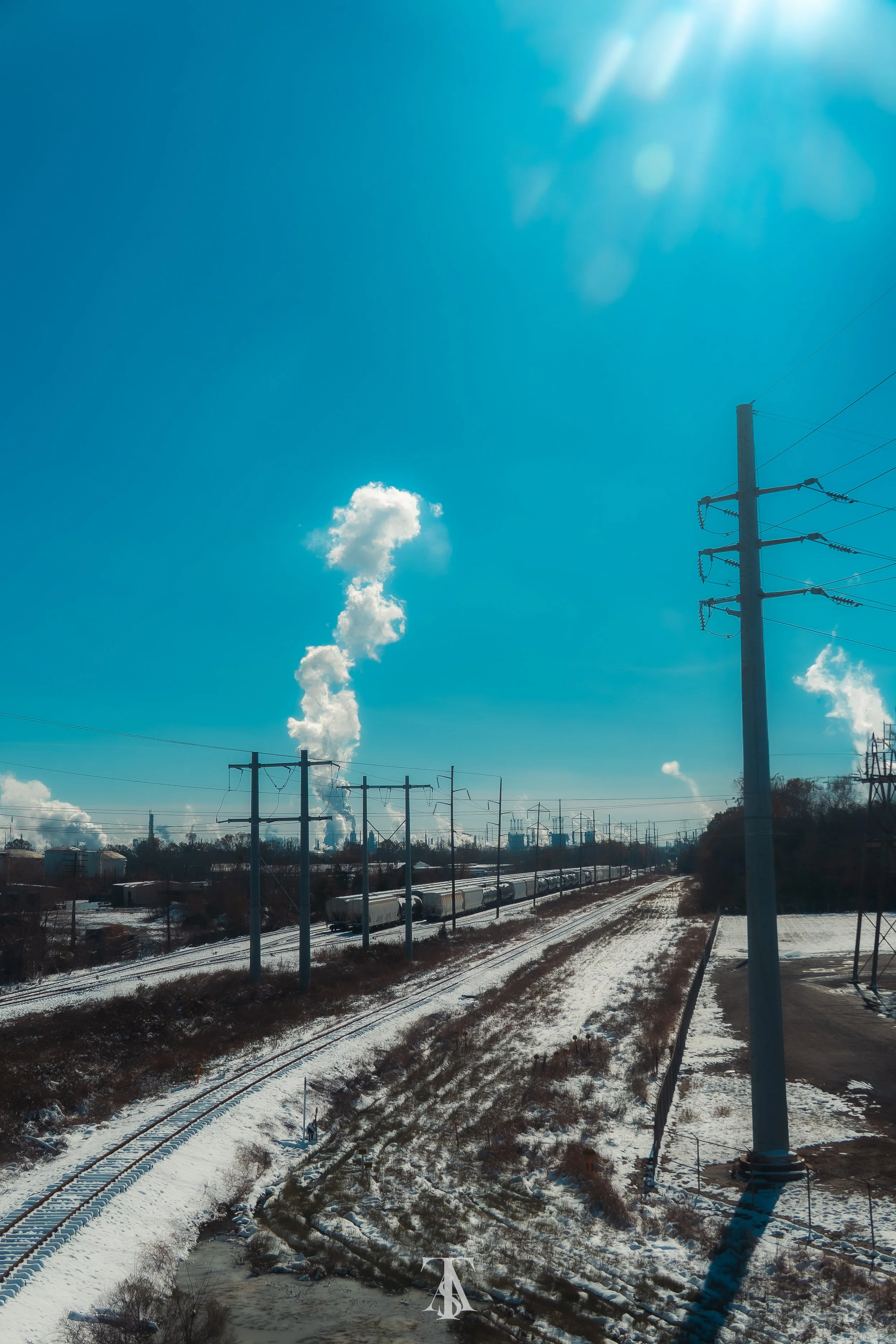 A train moving through a snow-covered landscape under a bright clear blue sky with the sun shining. Smoke or steam releases from factory chimneys in the distant background. Power lines and utility poles run alongside the train tracks.