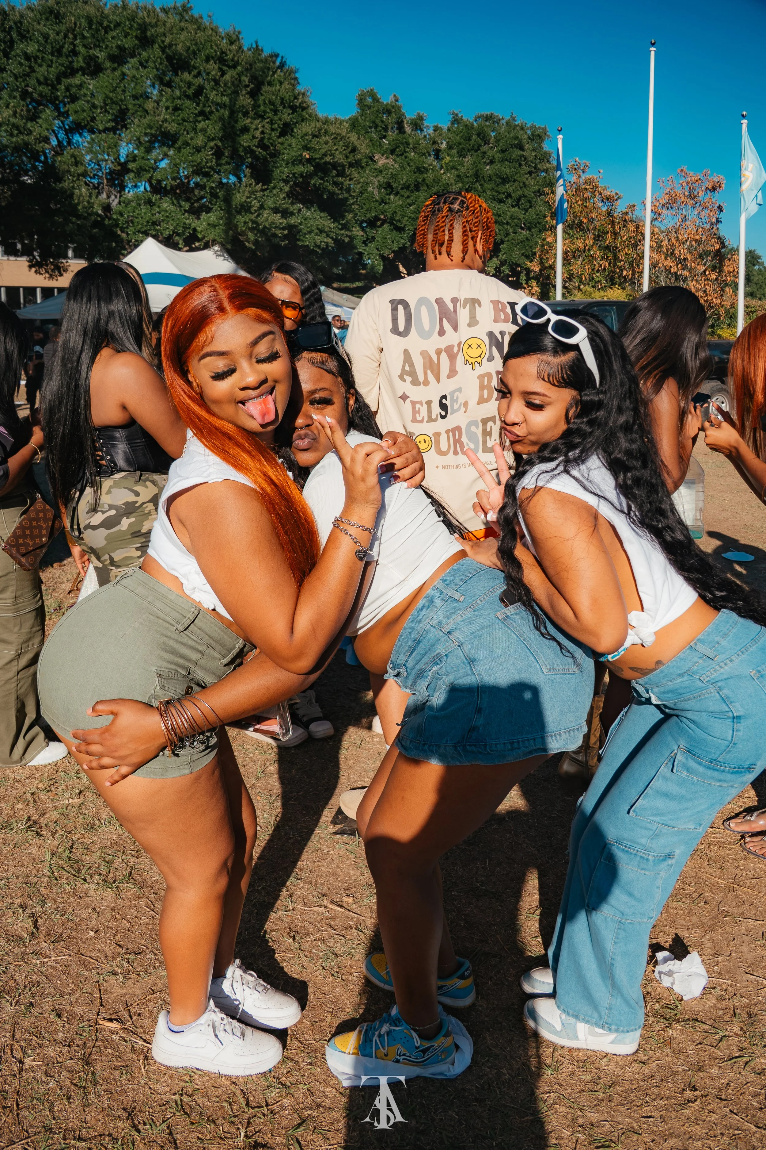 Three young women pose together outdoors at a gathering, making playful gestures and smiling, with a group of people and trees in the background.