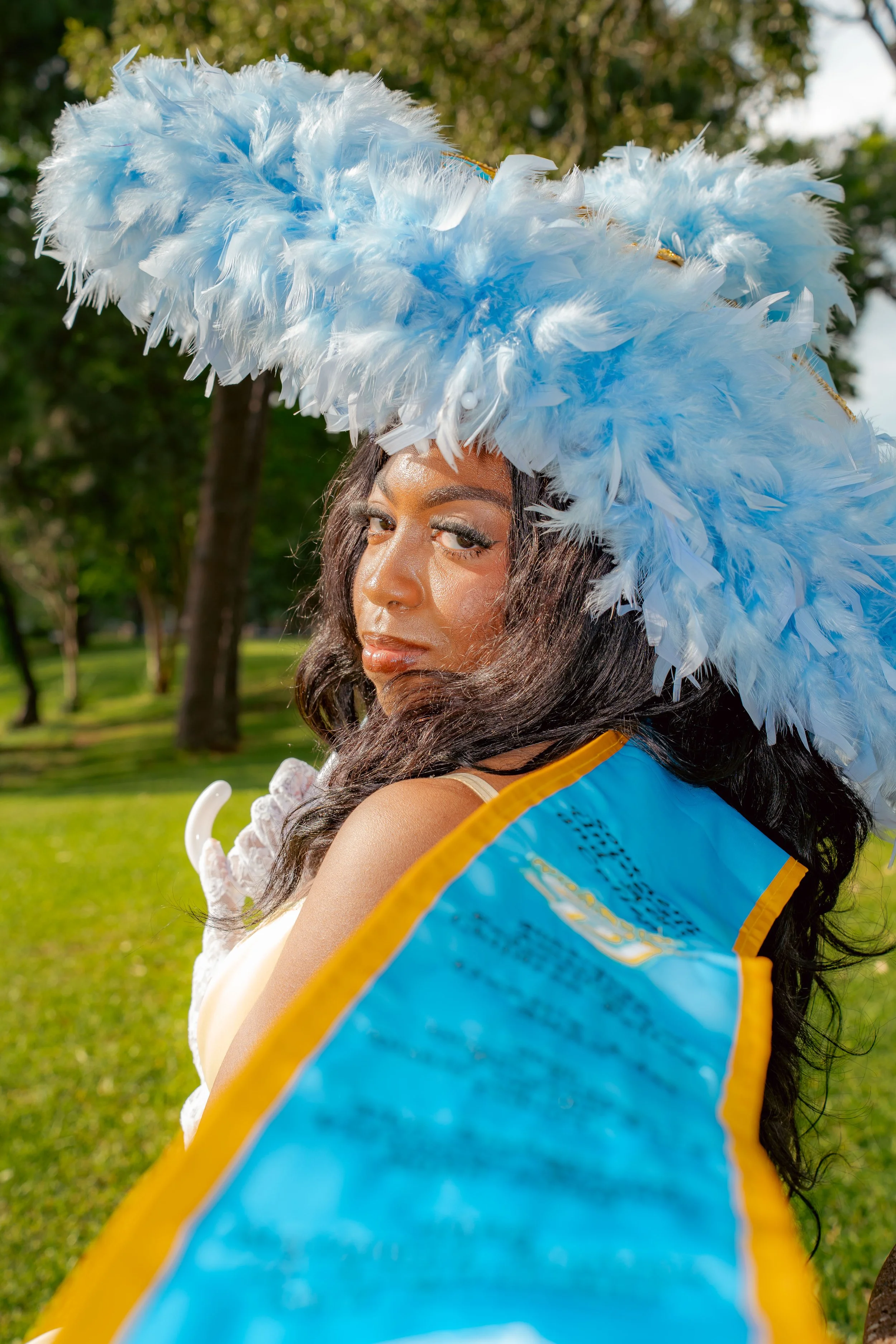 A woman dressed in a costume with a large, elaborate blue feather headdress, outdoor setting with trees and grass in the background.