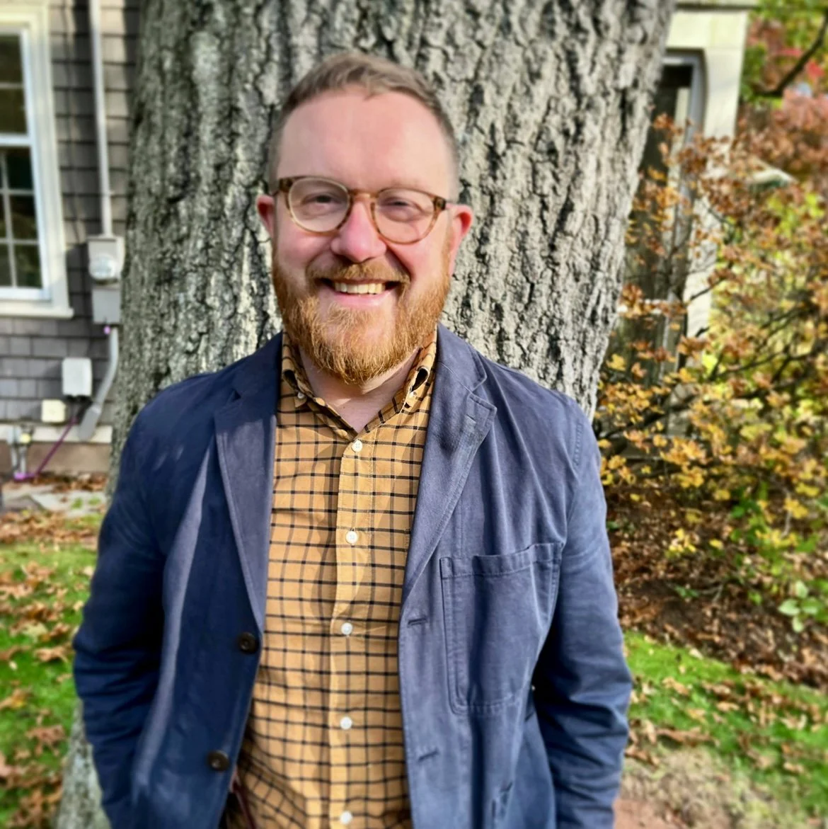 A person with glasses and a beard stands smiling against a large tree. The person is wearing a checkered shirt and a blue jacket. The background includes foliage and part of a building.