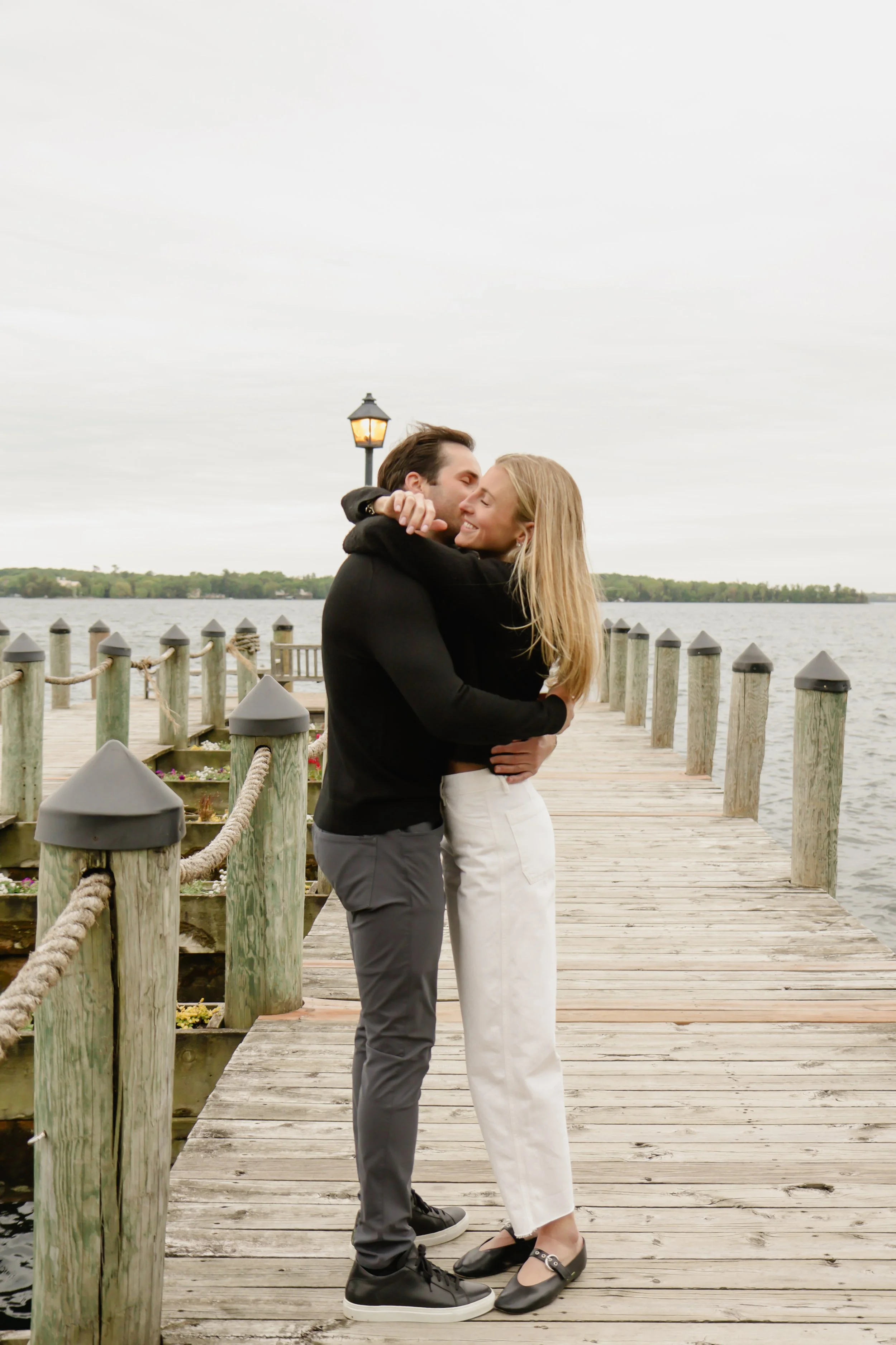 A couple hugging on a wooden pier with water and a shoreline in the background, overcast sky, lamp post nearby, both smiling