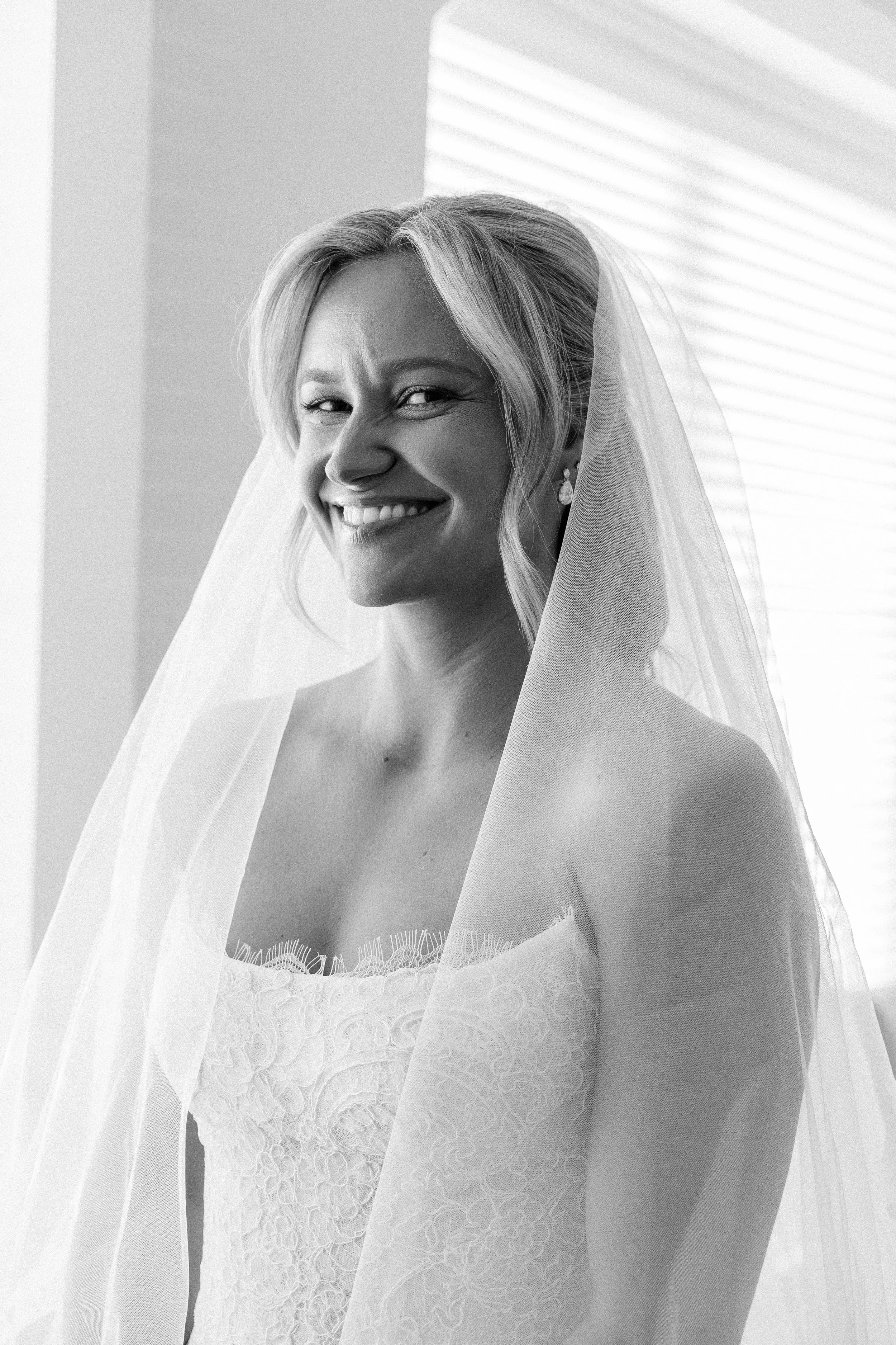 Black and white photo of a smiling bride with a veil and lace wedding dress, standing in front of a window with blinds.