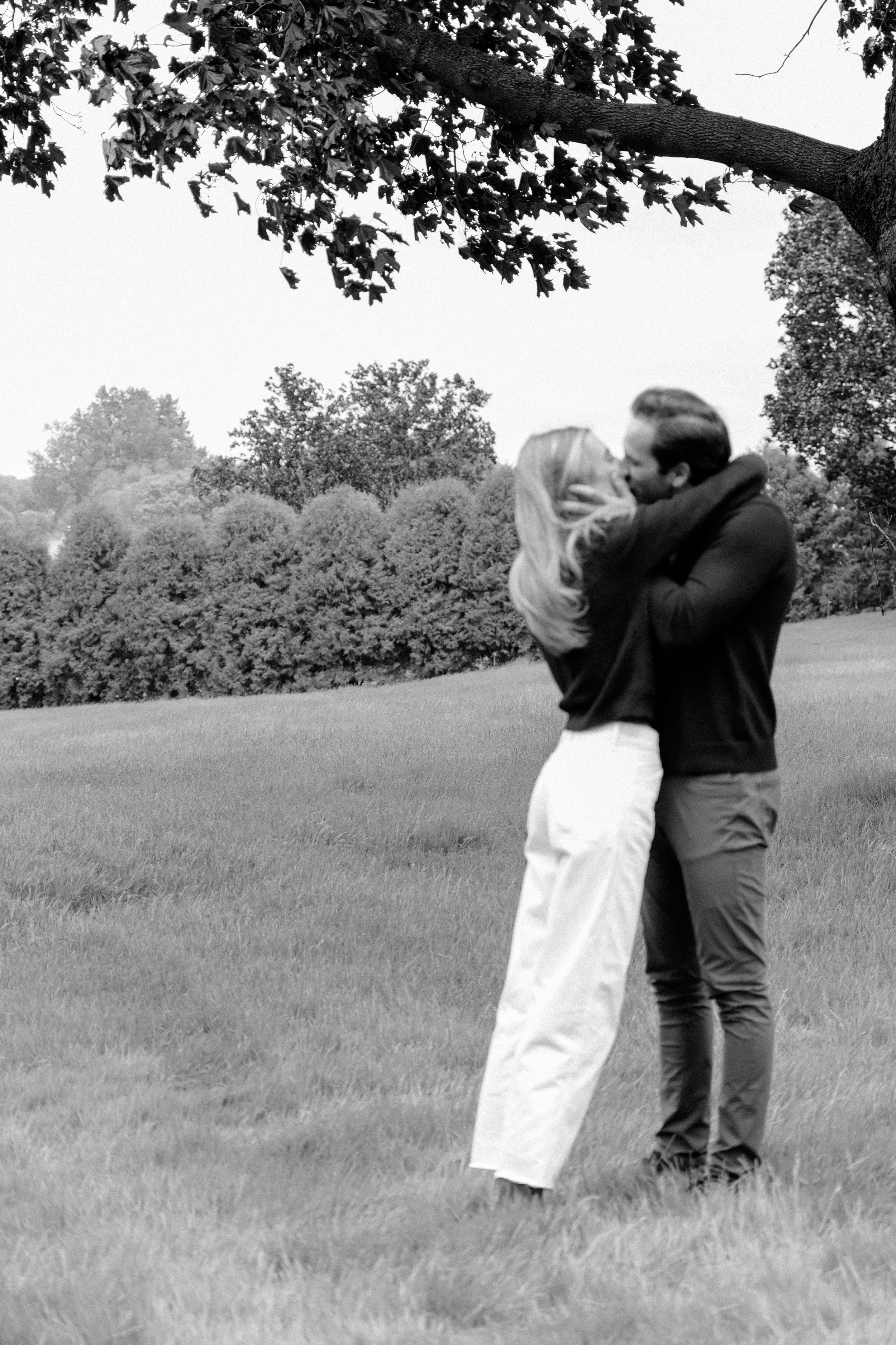 A black-and-white photograph of a couple kissing outdoors on a grassy field, with trees and shrubs in the background, and a large tree branch hanging overhead.
