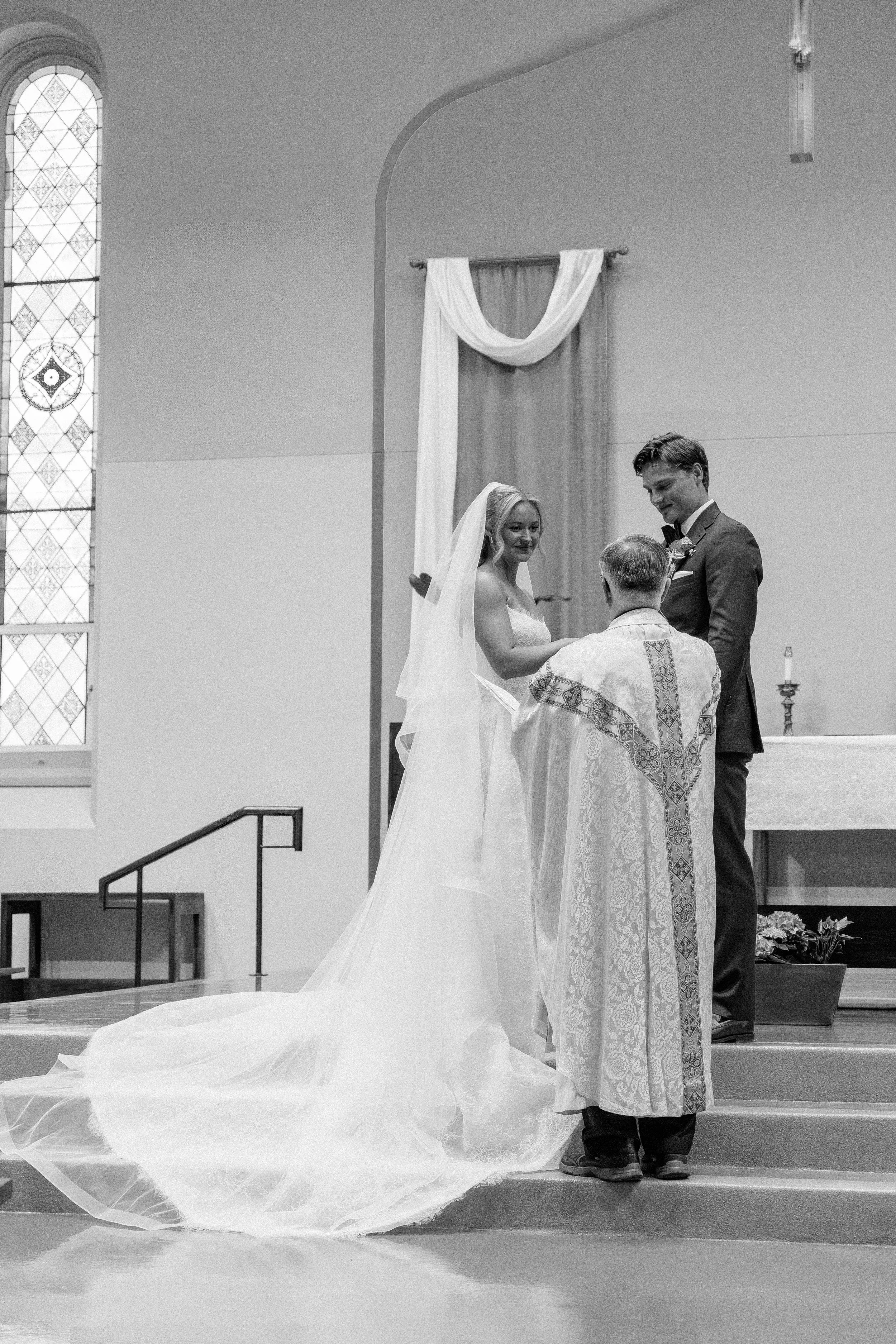 A bride and groom exchanging vows during a wedding ceremony in a church, with a priest officiating, a stained glass window on the left, and wedding decorations including draped fabric and flowers.