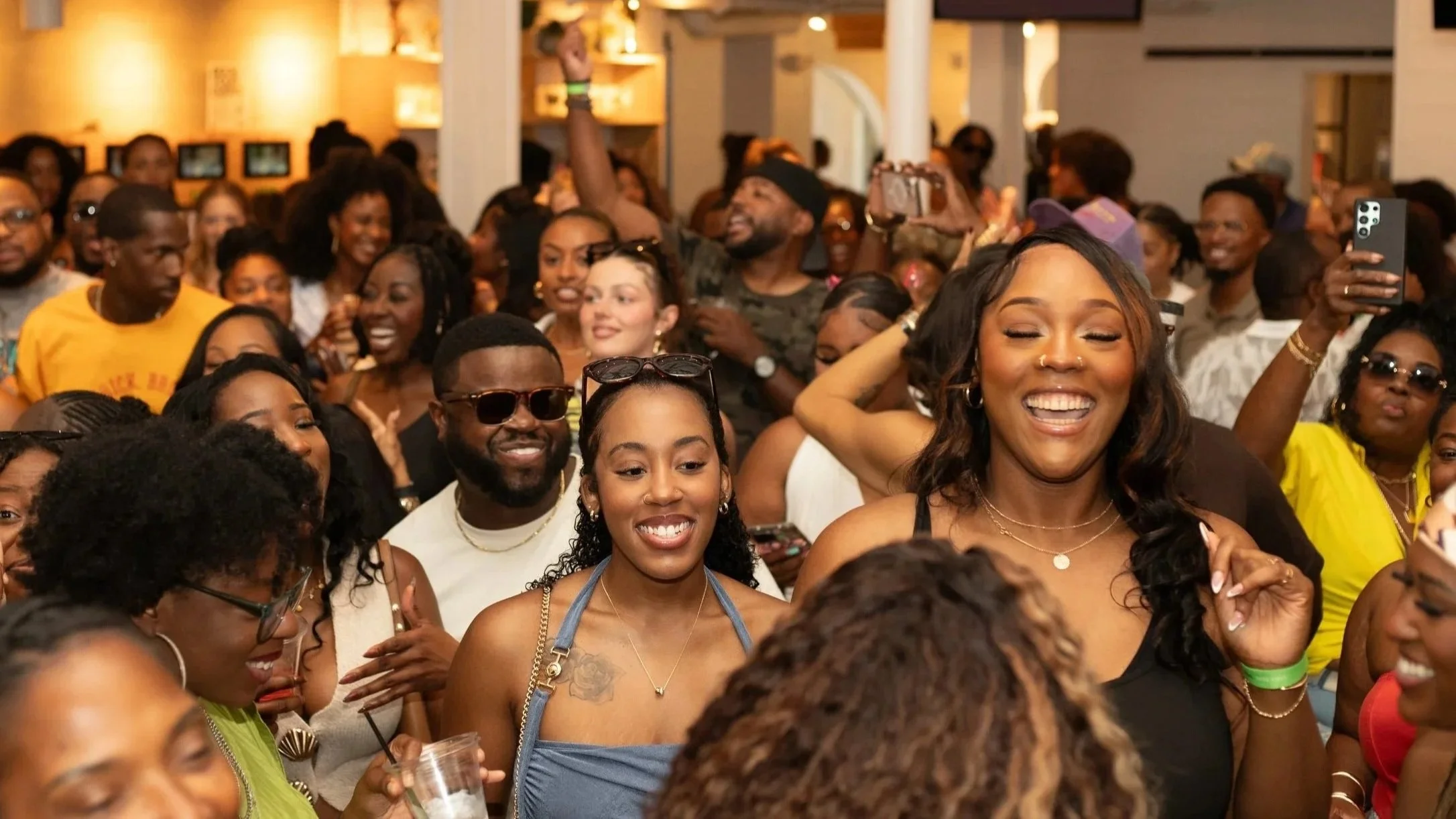 Large group of people dancing and socializing at an outdoor tennis court. The scene is lively with many individuals holding drinks, wearing casual summer attire, and some taking photos. A building in the background is painted teal, and the atmosphere appears festive.