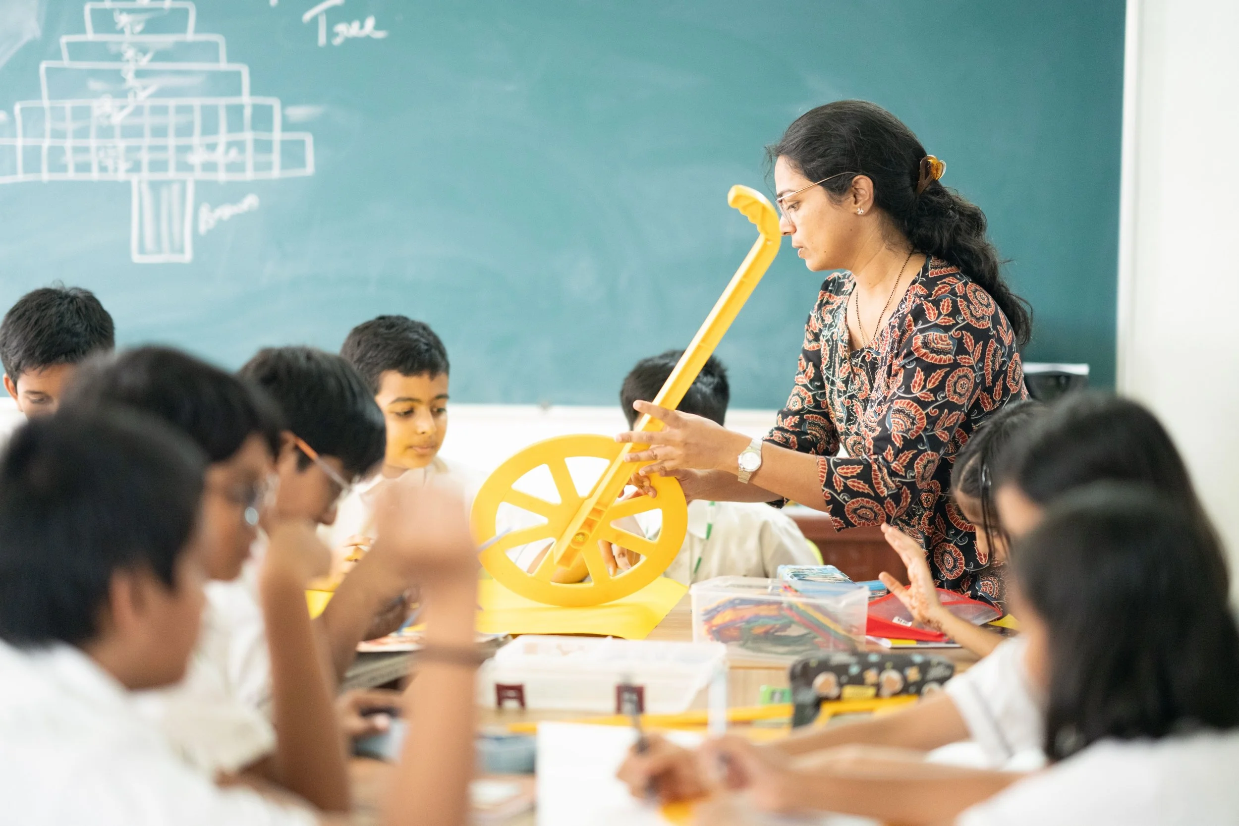 Teacher demonstrating a yellow measuring tool to students in a classroom with a chalkboard in the background.