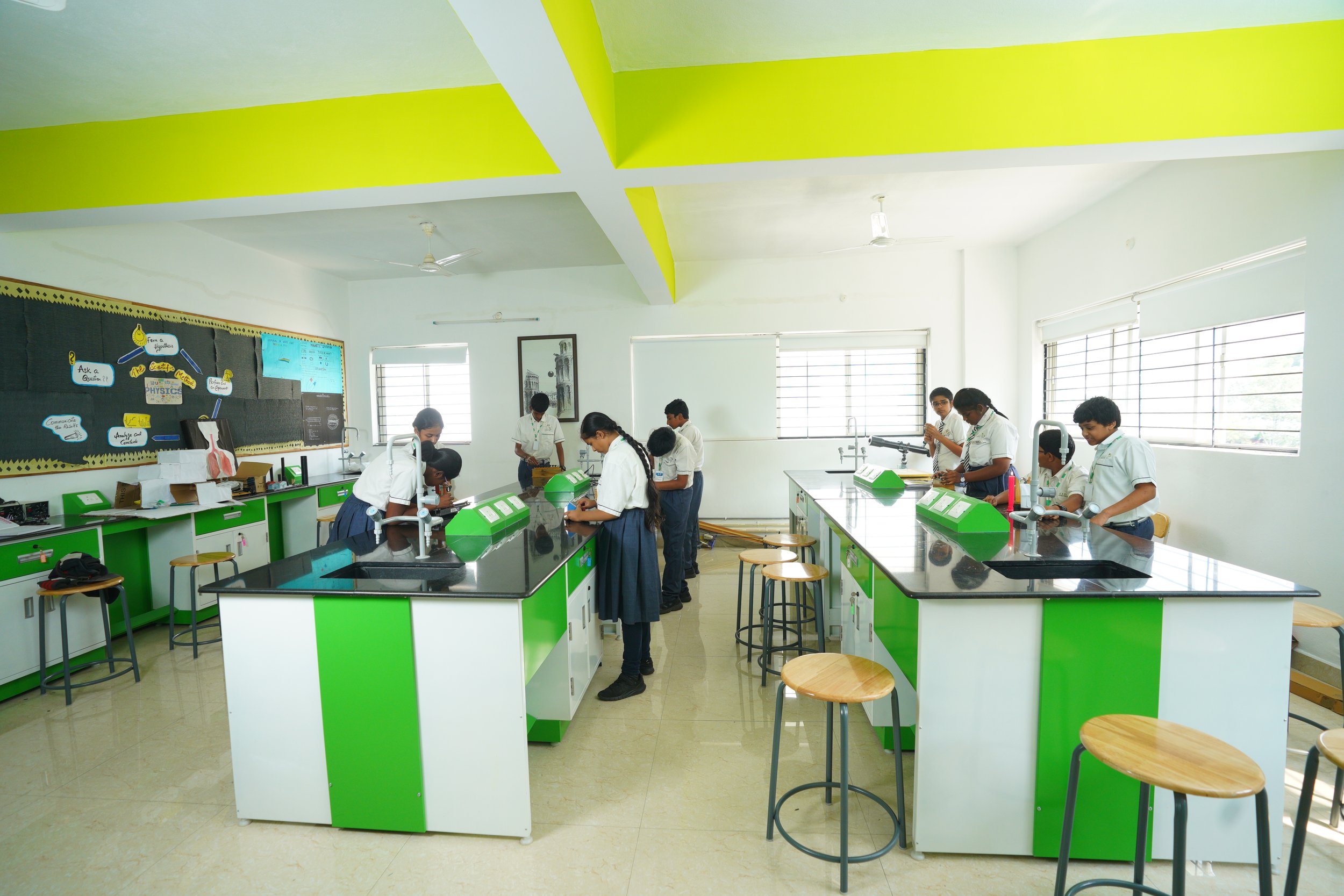 Students working in a bright science laboratory with green and white lab tables, stools, and experiment equipment. Large windows provide natural light and a bulletin board is visible in the background.