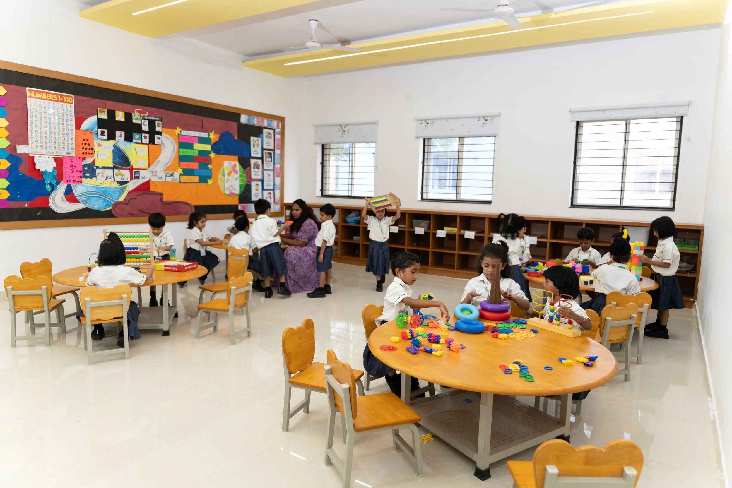 Preschool classroom with children engaged in various activities; tables with educational toys, a teacher assisting students, a colorful bulletin board, and wooden cubbies with supplies.