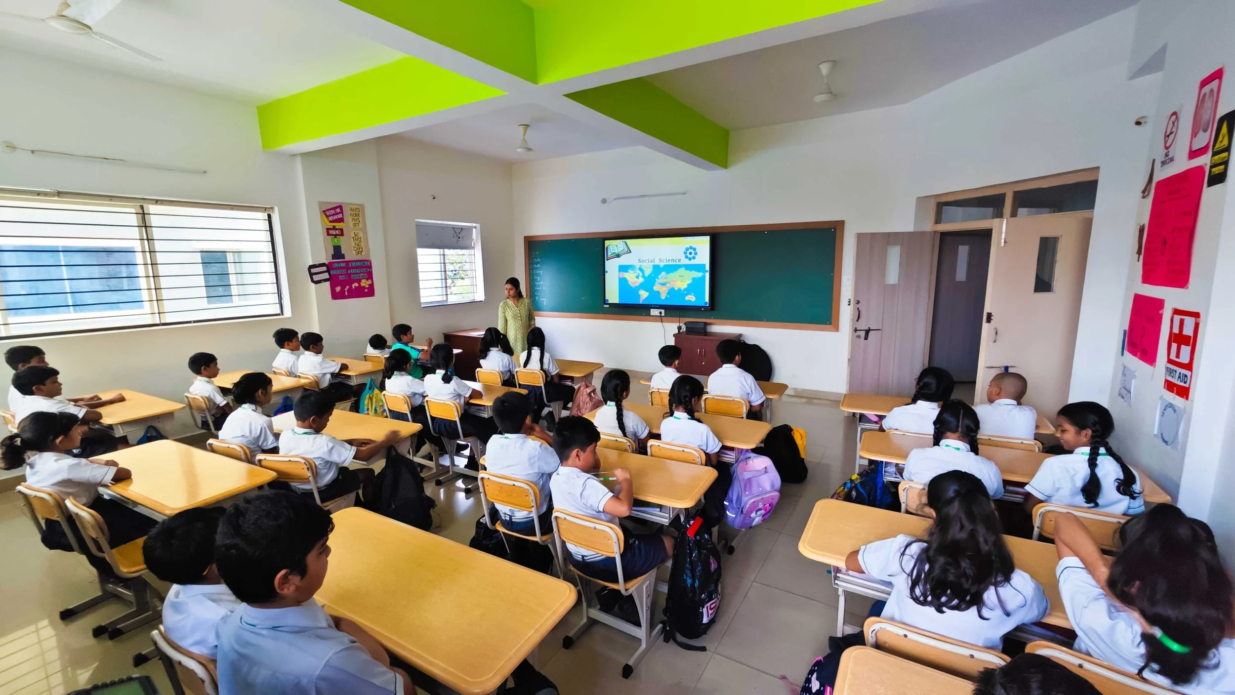 A classroom with students in uniforms sitting at desks, facing a teacher and a smart board displaying a world map under "Social Science." The classroom has bright green accents and posters on the wall.