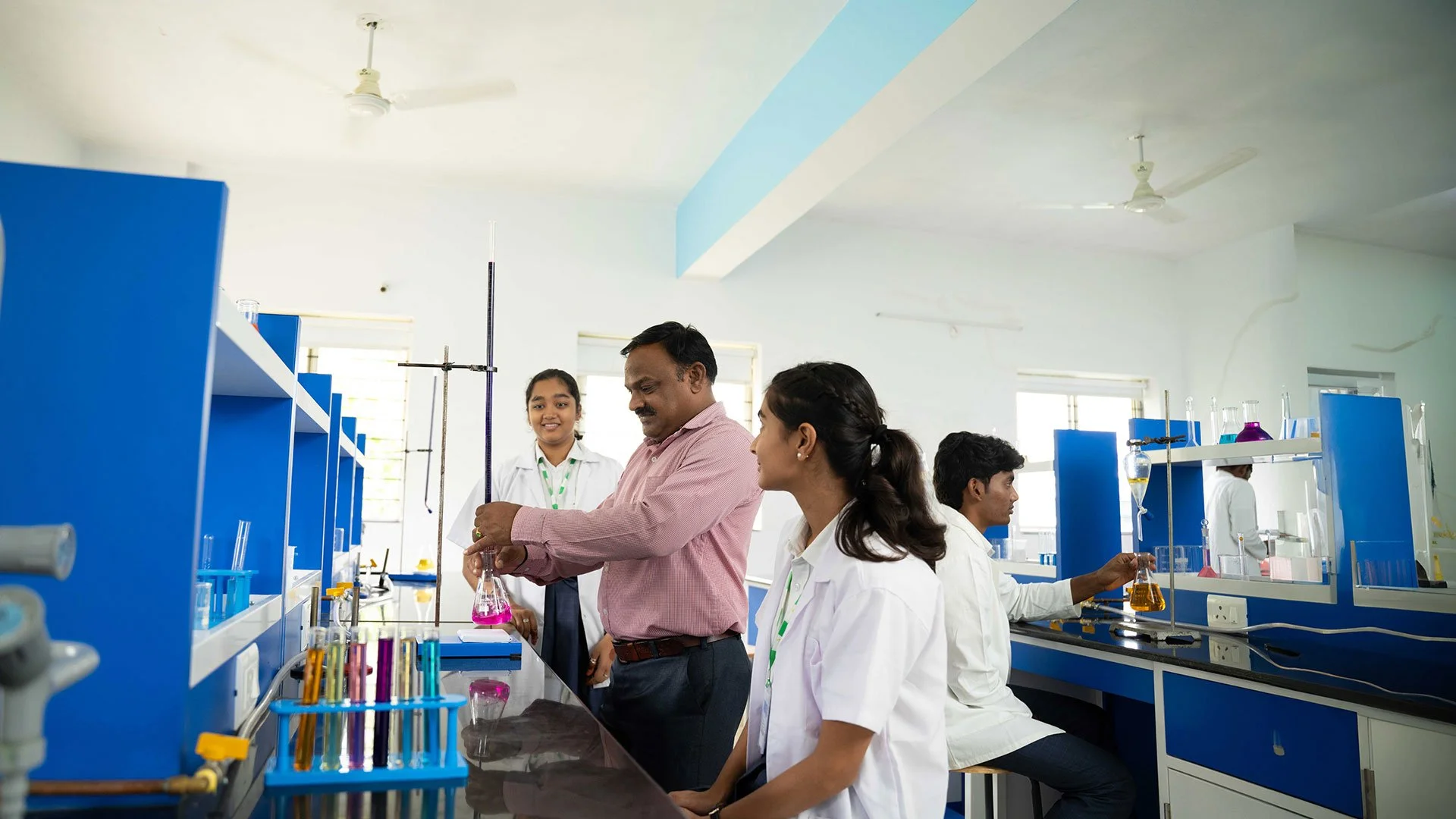 Students and a teacher conducting an experiment in a chemistry lab with blue workstations and lab equipment.