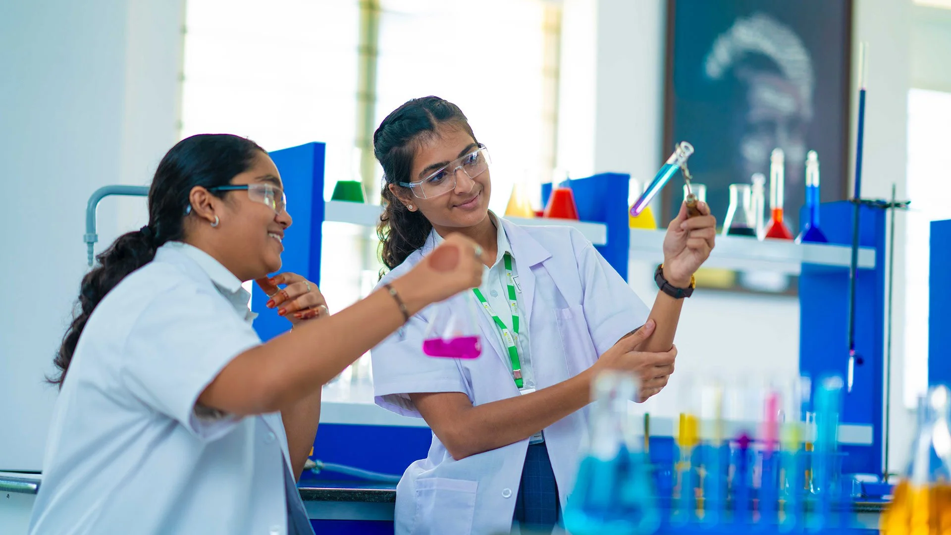 Two female students in a laboratory wearing lab coats and goggles, examining a test tube with colored liquid.