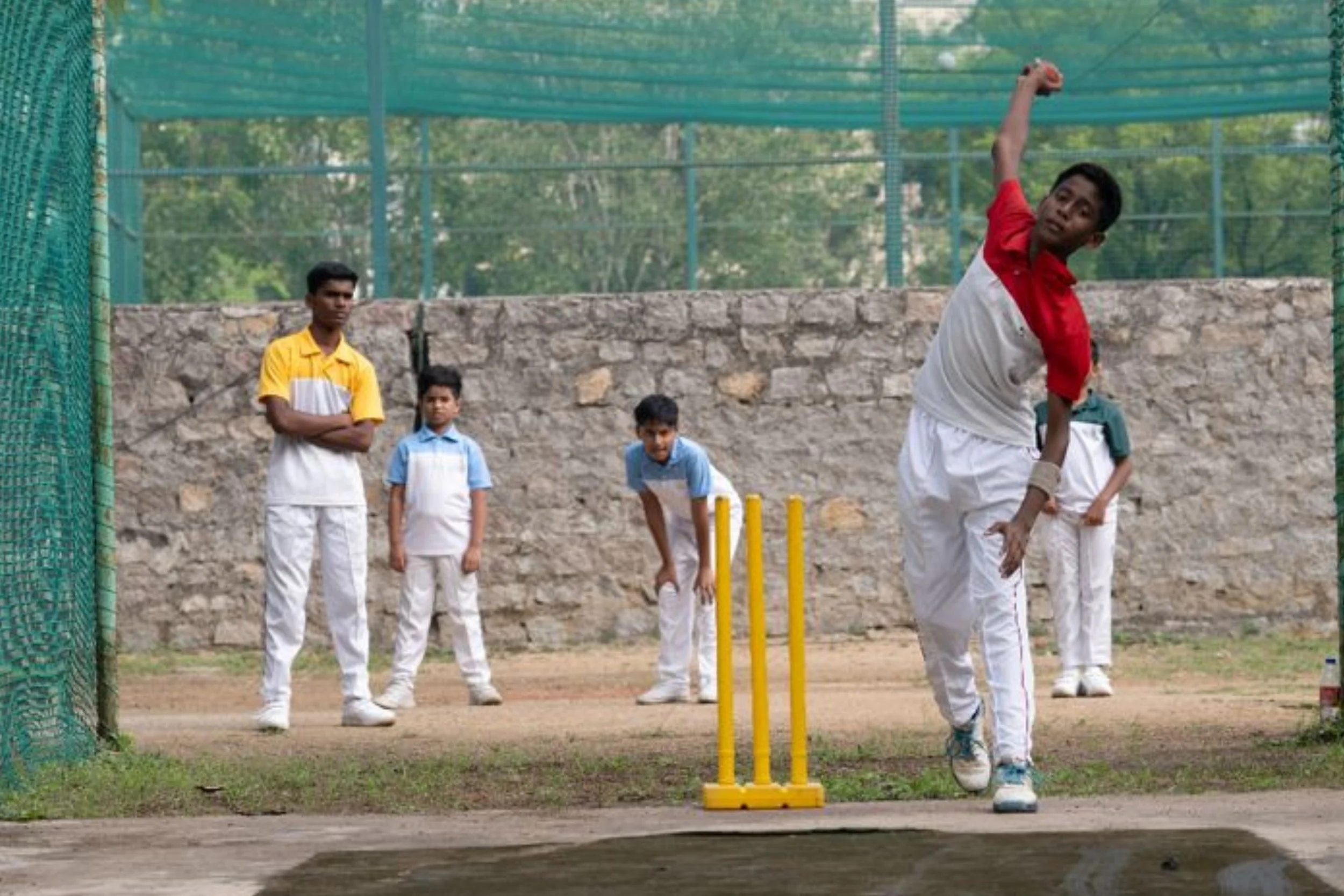 Group of boys playing cricket, one boy bowling, others watching, with wickets in the foreground and netting in the background.