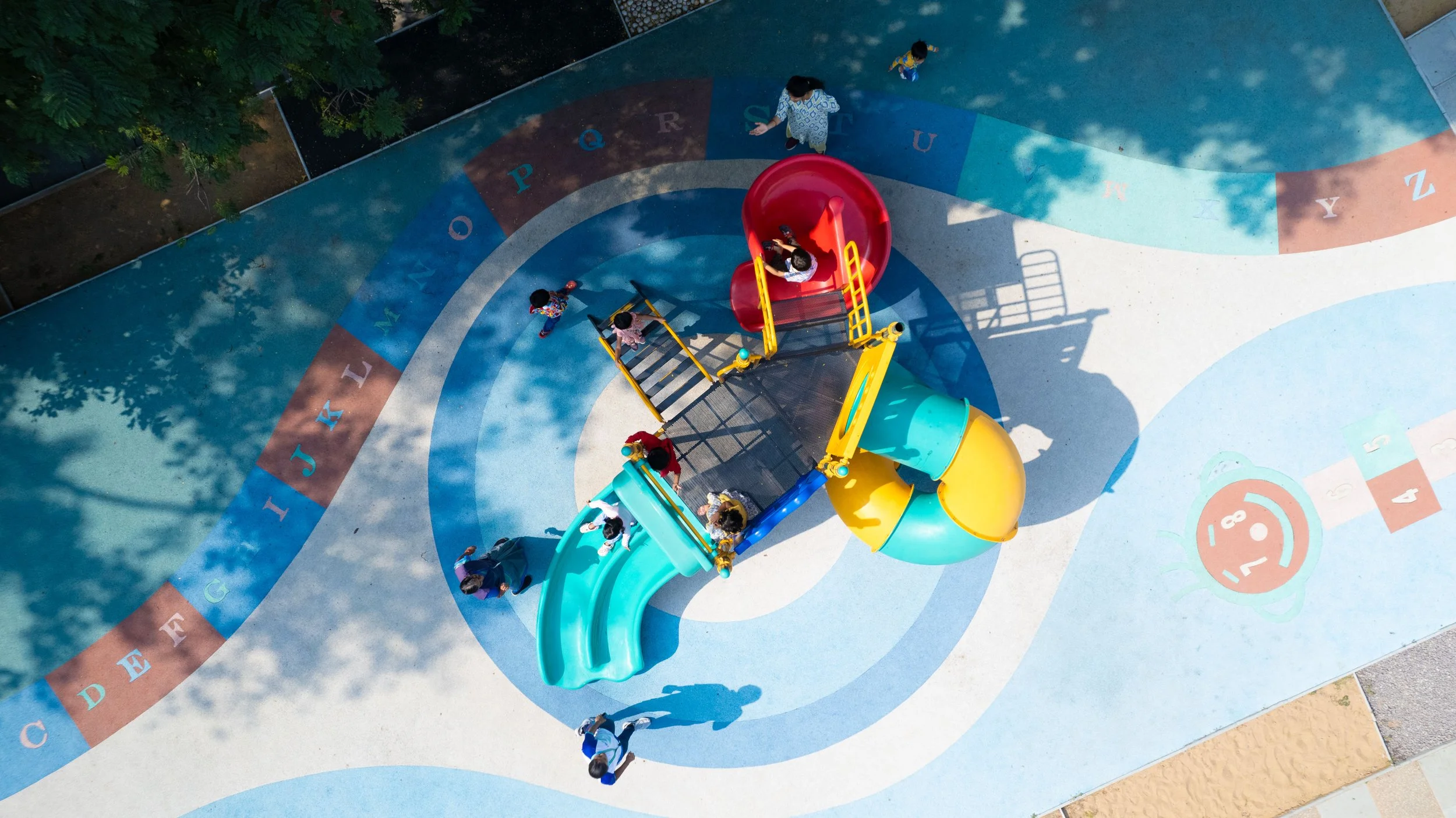 Aerial view of a colorful playground with slides and children playing. The ground features concentric circles and alphabet letters in a playful design.