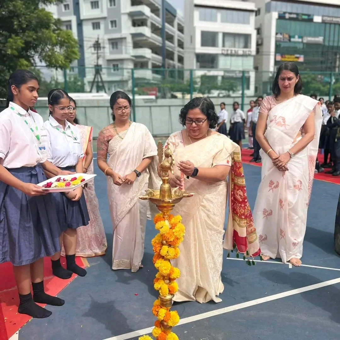 A group of women and students participating in a traditional Indian lamp lighting ceremony on an outdoor basketball court, with multi-story buildings in the background.