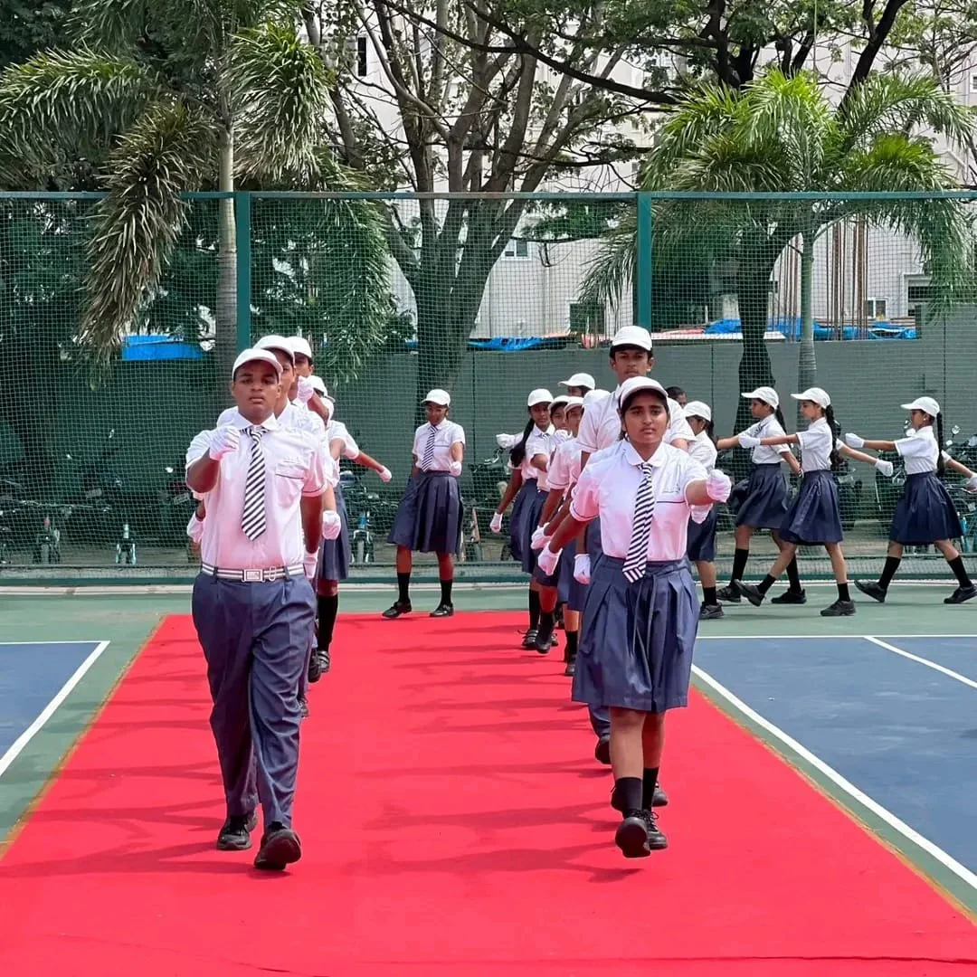 School children in uniform walking on a red carpet on a tennis court, with some saluting and others dancing in the background.