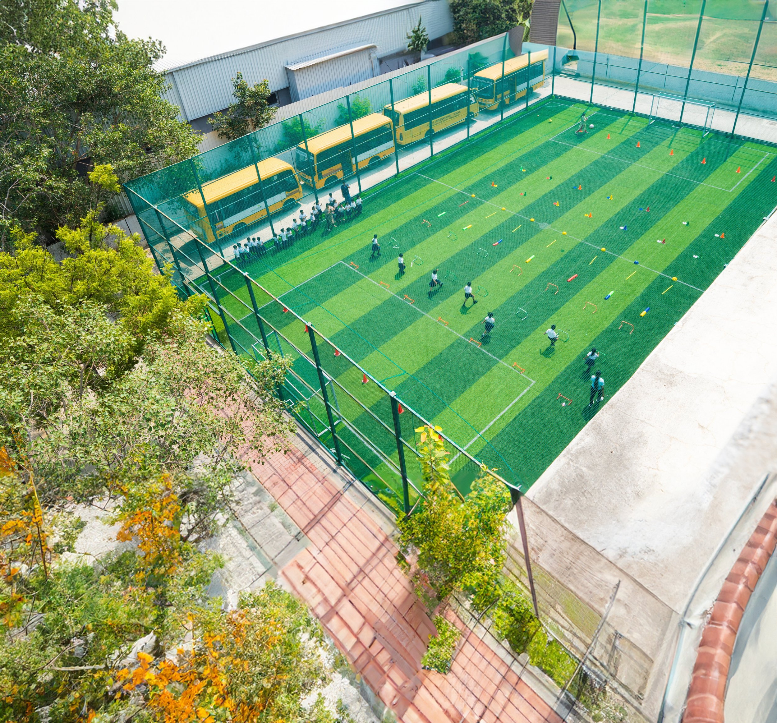Aerial view of a fenced sports field with children playing. The field has artificial green turf and is surrounded by netting. Yellow school buses are parked beside the field. Trees and a paved path are visible nearby.