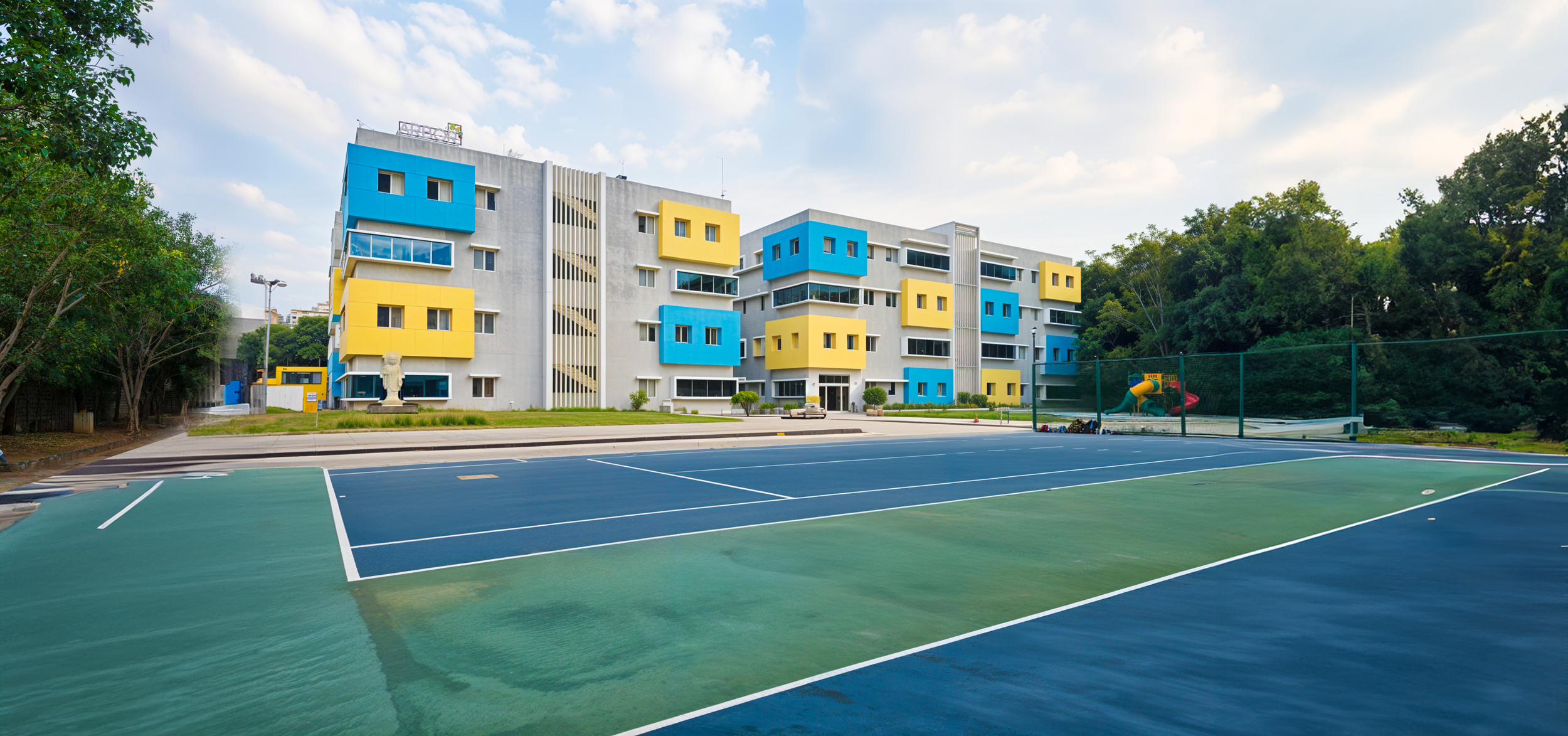 Colorful school building with blue and yellow sections next to a tennis court, surrounded by trees.