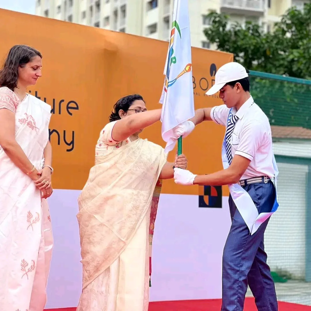A woman in a cream saree is receiving a flag from a young man in a school uniform and cap, during a ceremony on an outdoor stage, with another woman in a saree watching nearby.