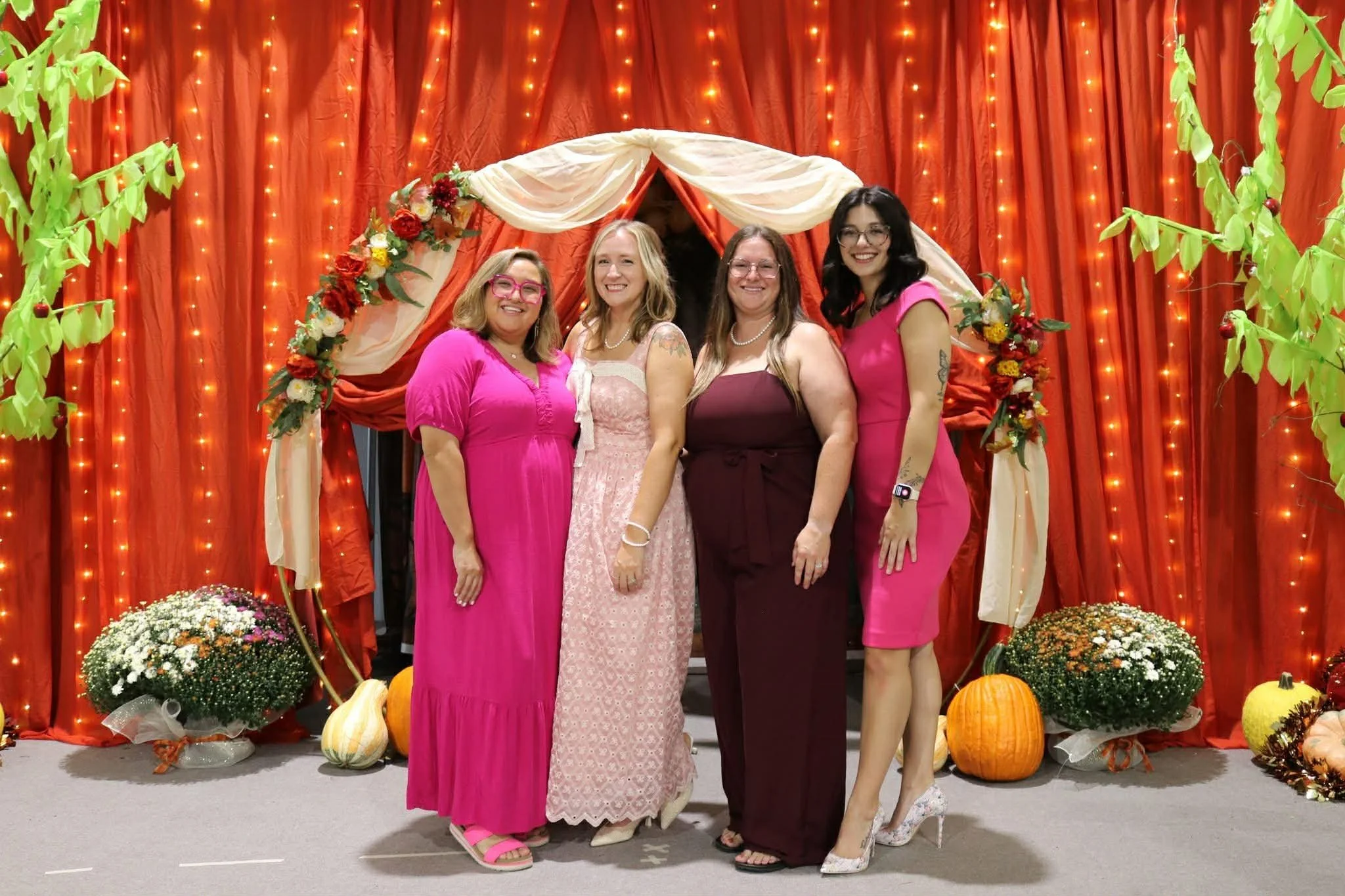 Four women standing together at an indoor event with orange curtains, string lights, flowers, pumpkins, and paper trees in the background. All women are smiling and wearing pink or purple dresses.