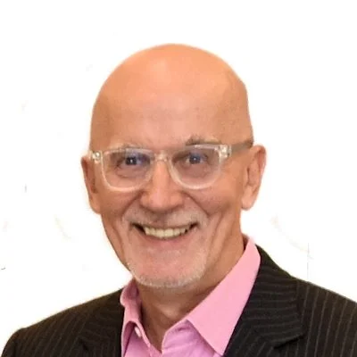 A smiling elderly man with glasses, bald head, wearing a pink shirt and black pinstripe suit
