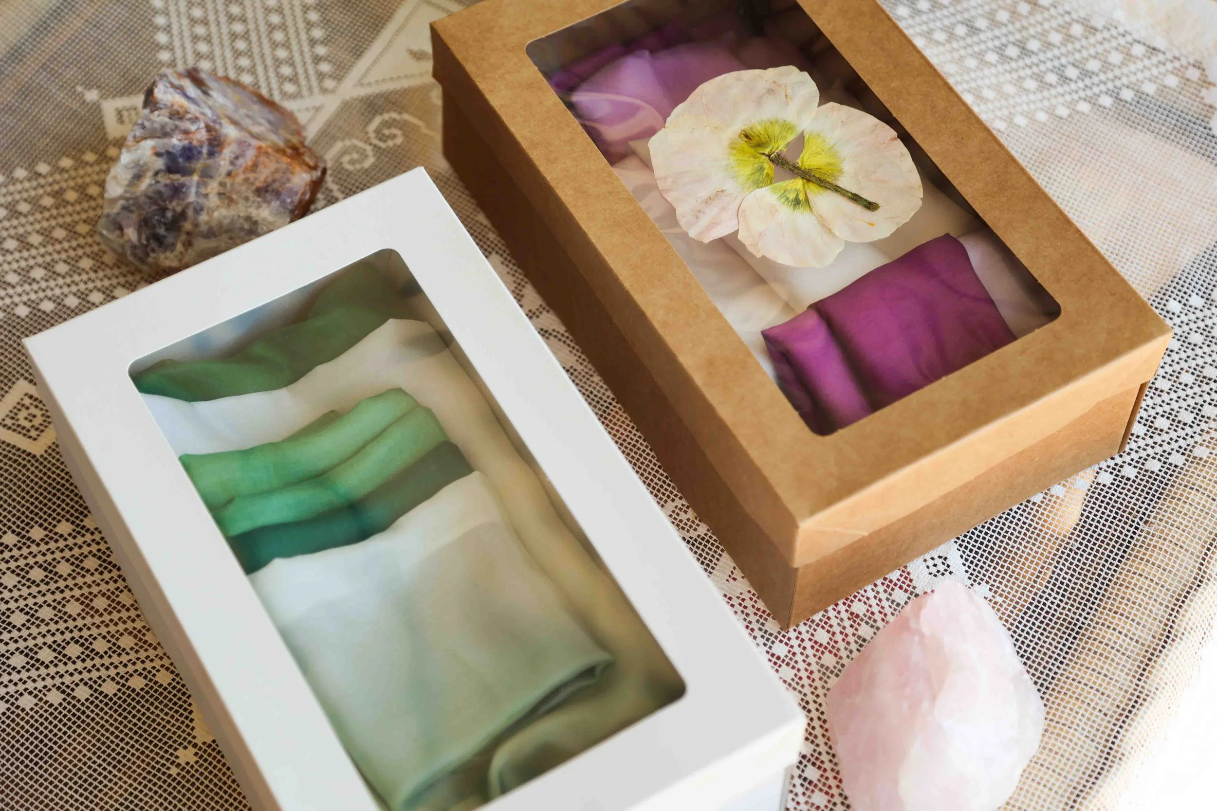 Two boxes containing folded scarves in various colors, one with a clear window, placed on a patterned cloth. Decorative stones are nearby.