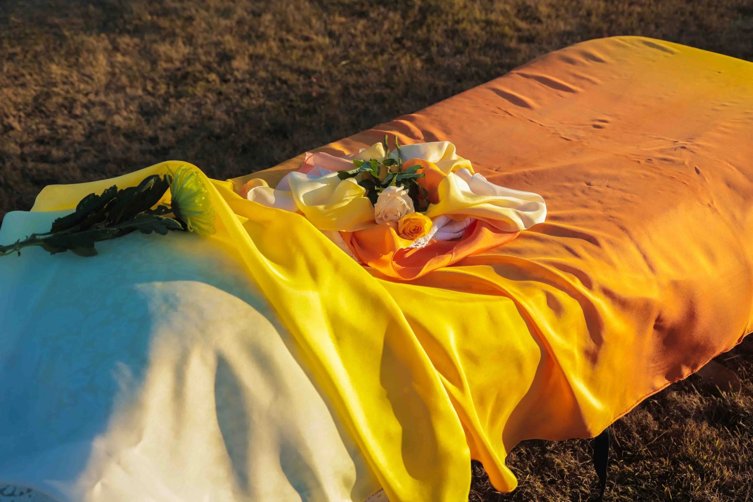 A table covered with orange and yellow fabric, adorned with white and yellow roses, on a grassy outdoor setting.
