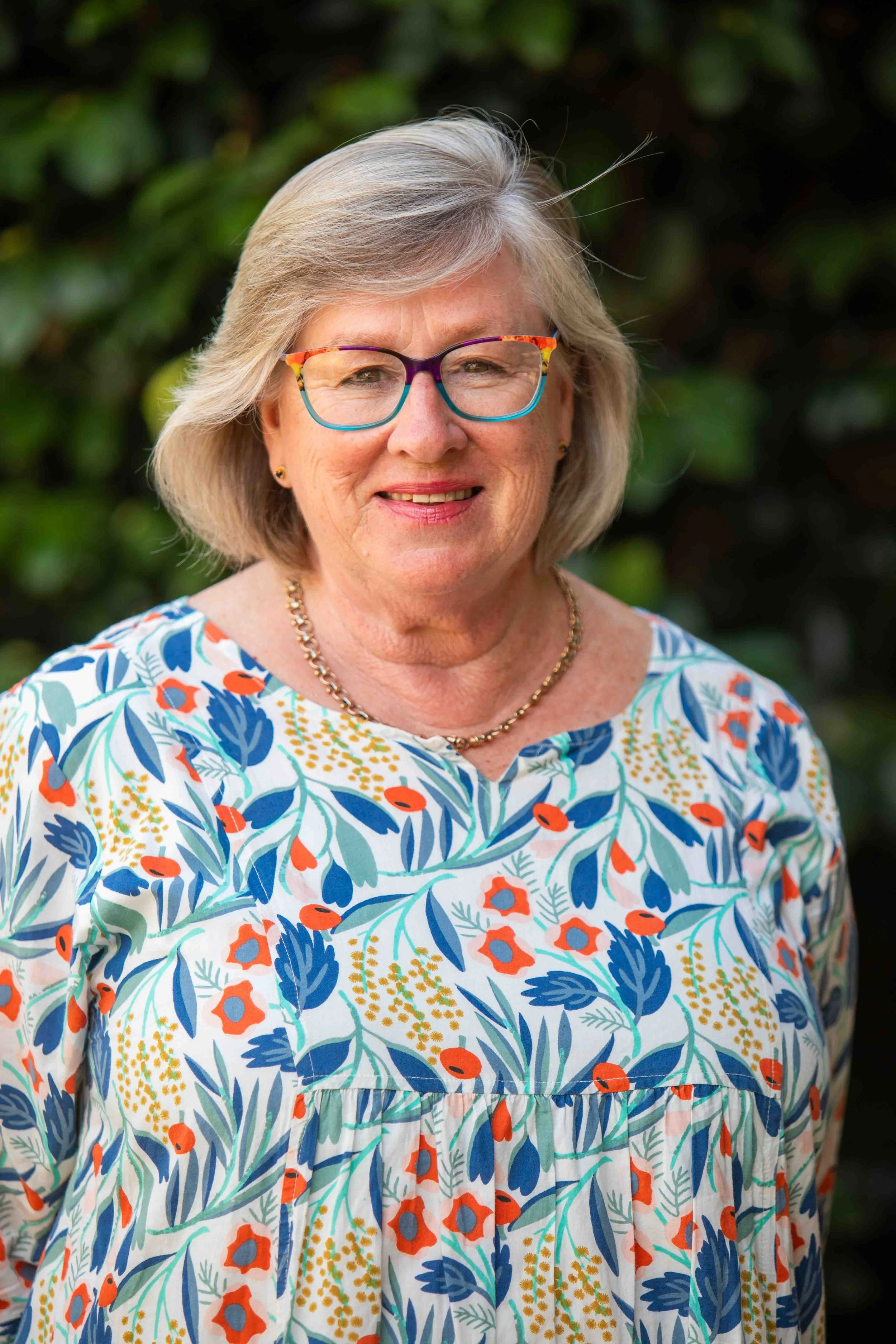 Elderly woman with gray hair wearing colorful glasses and a floral dress, standing outdoors.