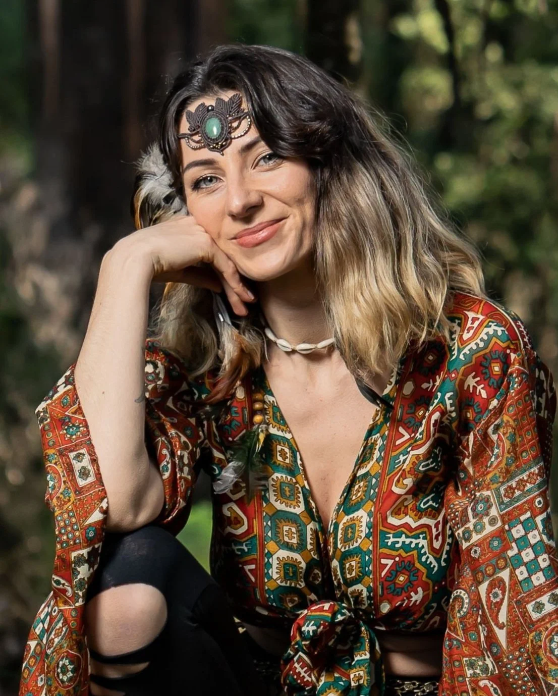 A woman sitting outdoors, wearing a colorful patterned top and a headpiece with a green stone. She is smiling and resting her hand on her chin. The background is blurred greenery.