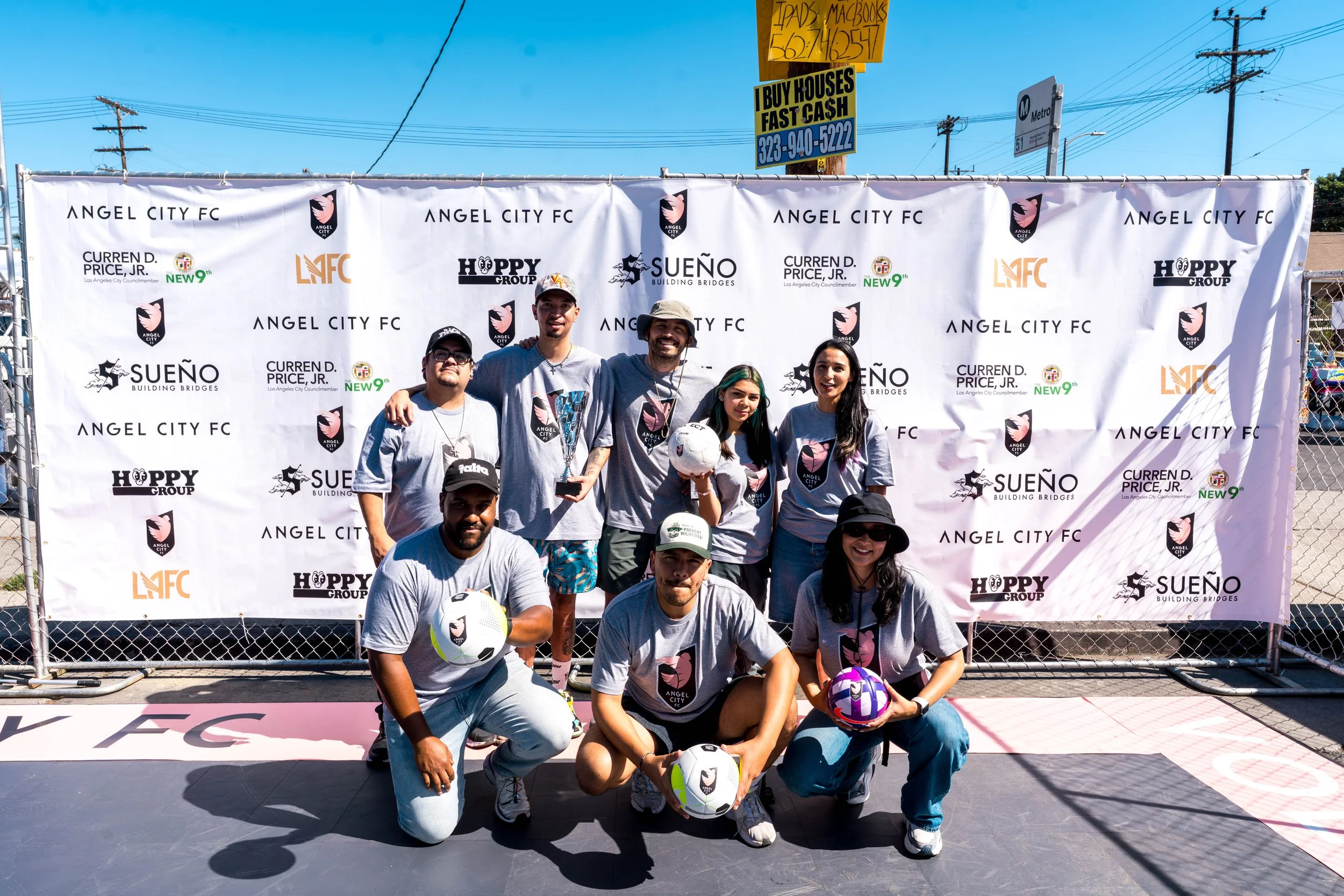 A group of nine people pose in front of a backdrop with the Angel City FC logo and sponsor logos, holding soccer balls, on an outdoor court on a sunny day.