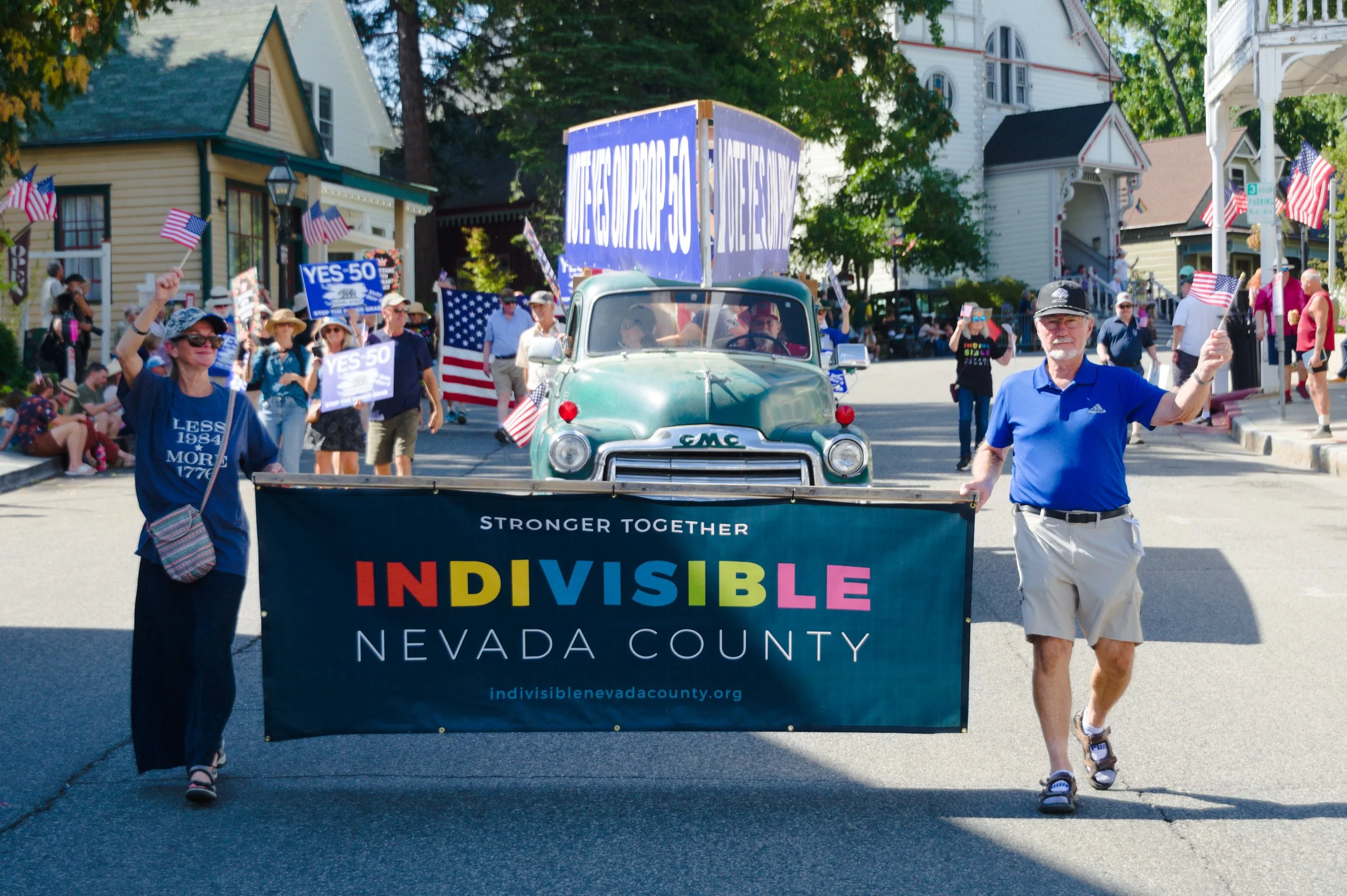 People participating in a parade, holding a banner for Nevada County, with a vintage GMC truck behind, decorated with signs and flags supporting political campaigns, in a neighborhood with houses and trees.