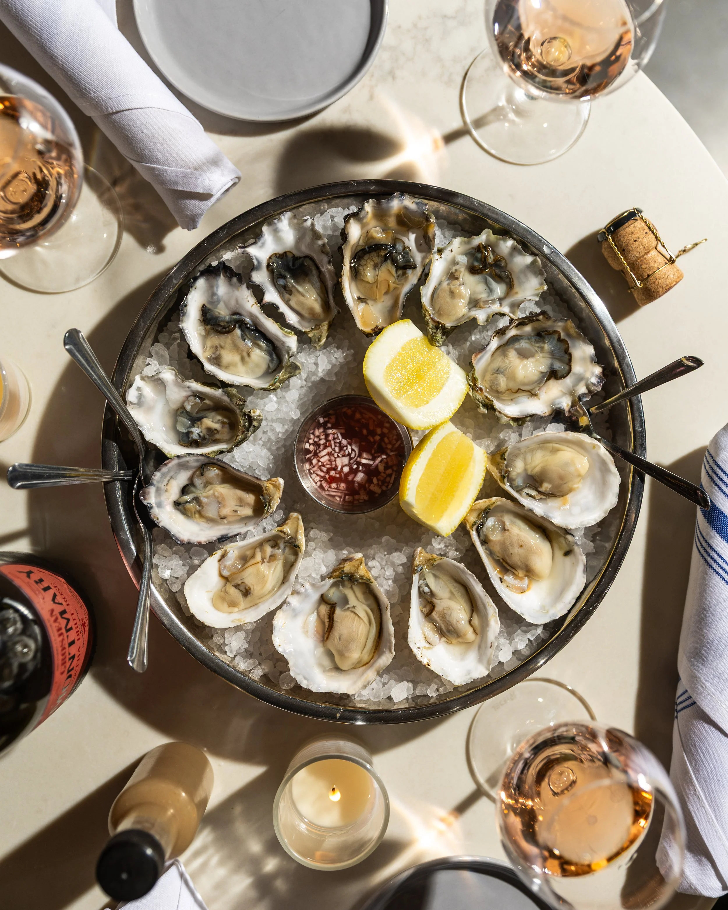 A round tray of fresh oysters on ice with lemon wedges and cocktail sauce in the center, surrounded by glasses of rosé wine, a candle, and a bottle of wine on a white table.