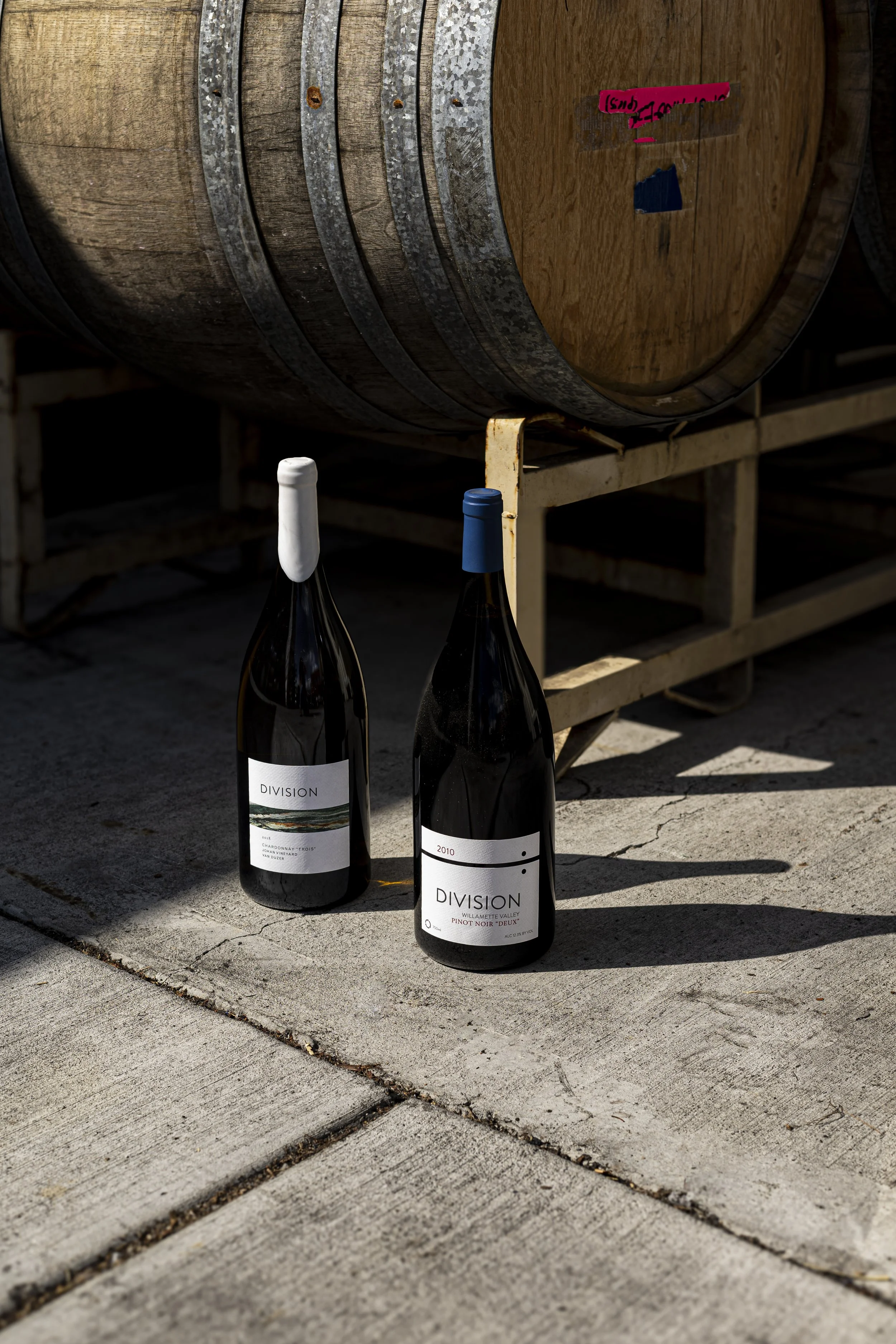 Two bottles of wine placed on a concrete sidewalk next to a large wooden wine barrel with metal bands, outdoors in sunlight.