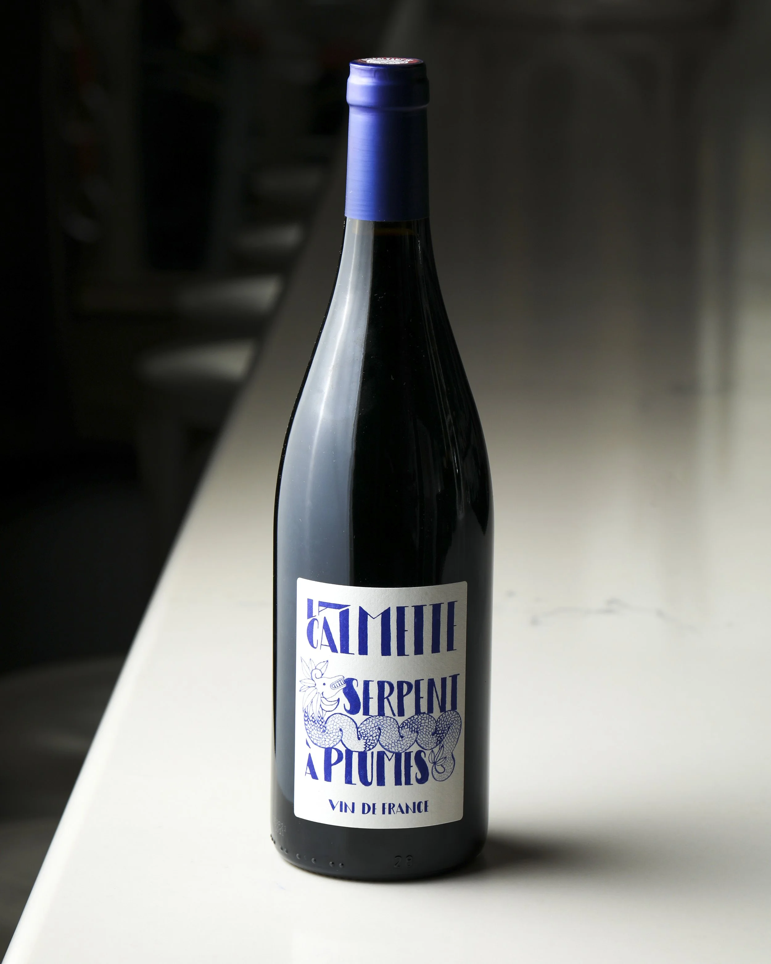 A dark bottle of wine with a white label and blue cap, standing on a white surface with a dark background.