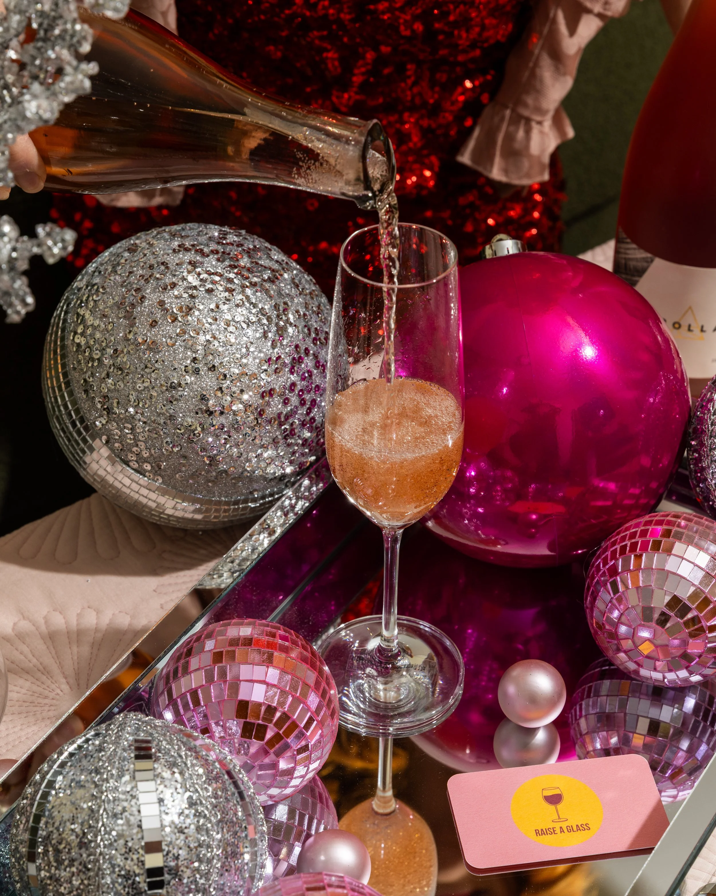 A glass of bubbly pink beverage being poured, surrounded by pink and silver Christmas ornaments on a reflective surface with a pink card reading 'Raise a Glass'.