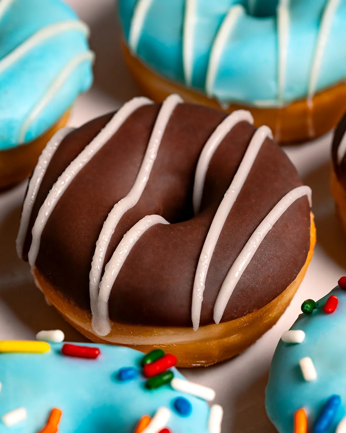 Close-up of chocolate-glazed donut with white icing drizzle, surrounded by blue frosted donuts with colorful sprinkles.