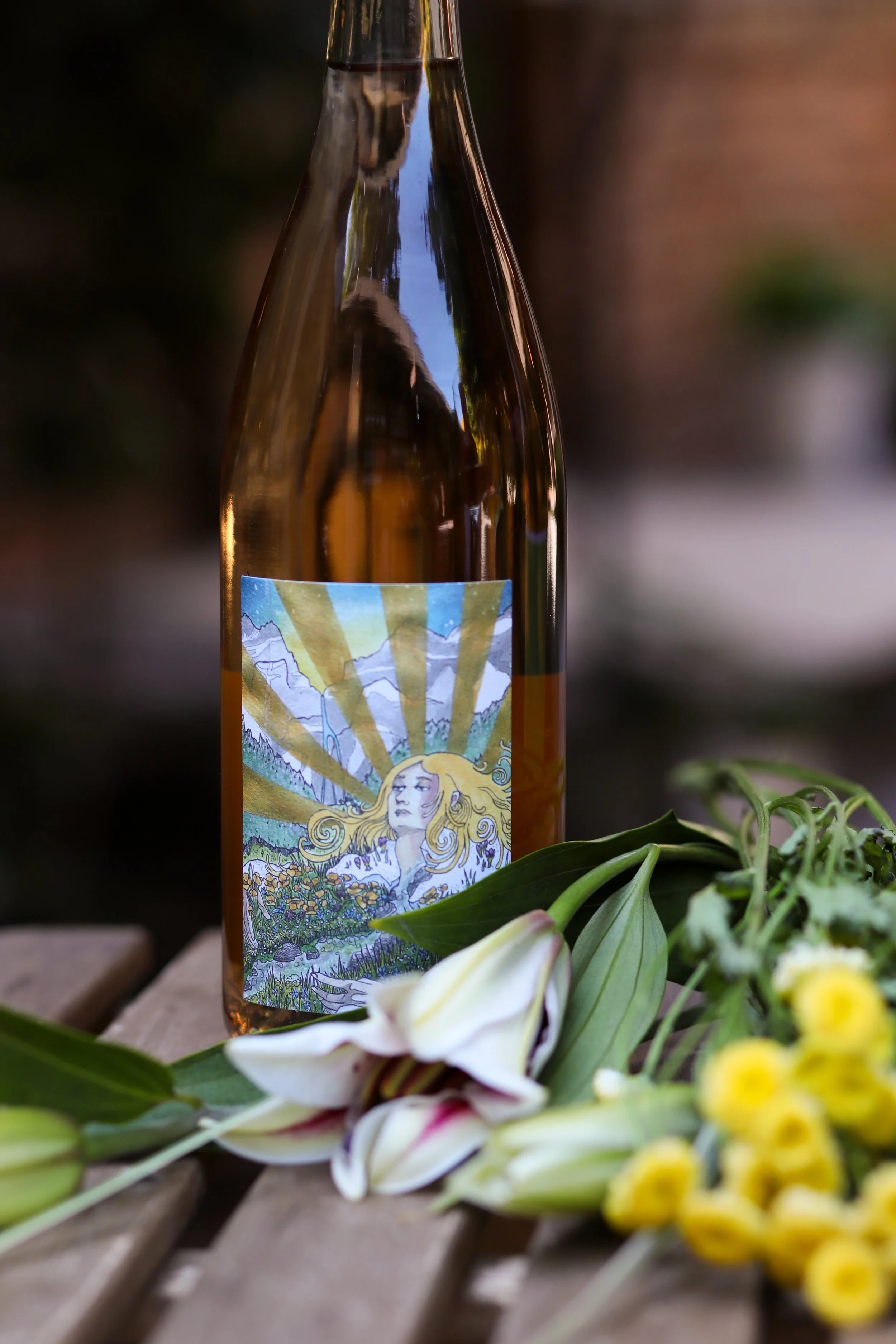 A brown glass bottle with a colorful label featuring a woman with flowing blonde hair, set outdoors on a wooden surface surrounded by white and yellow flowers.