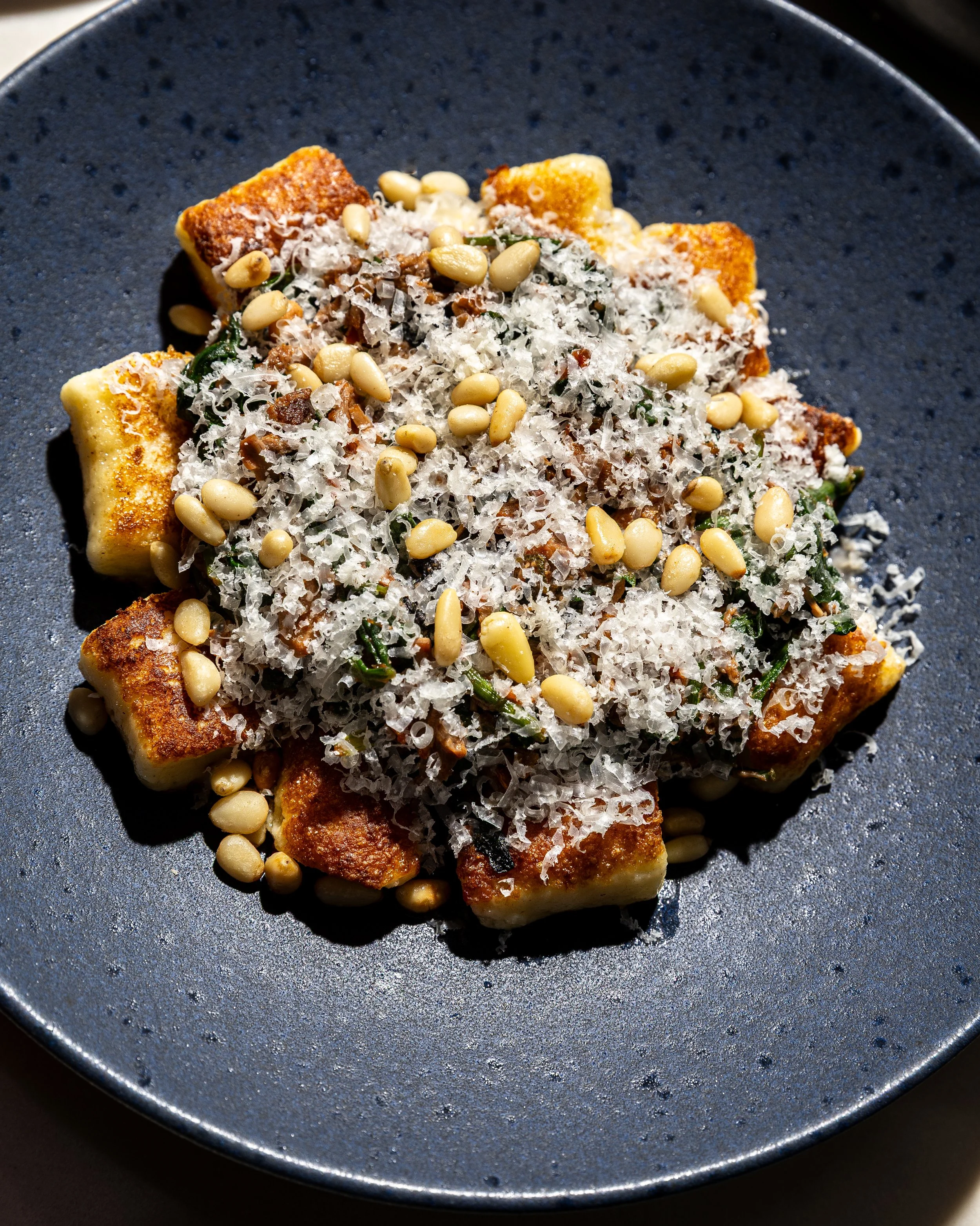 Fried tofu topped with shredded cheese, pine nuts, and chopped herbs on a dark plate.