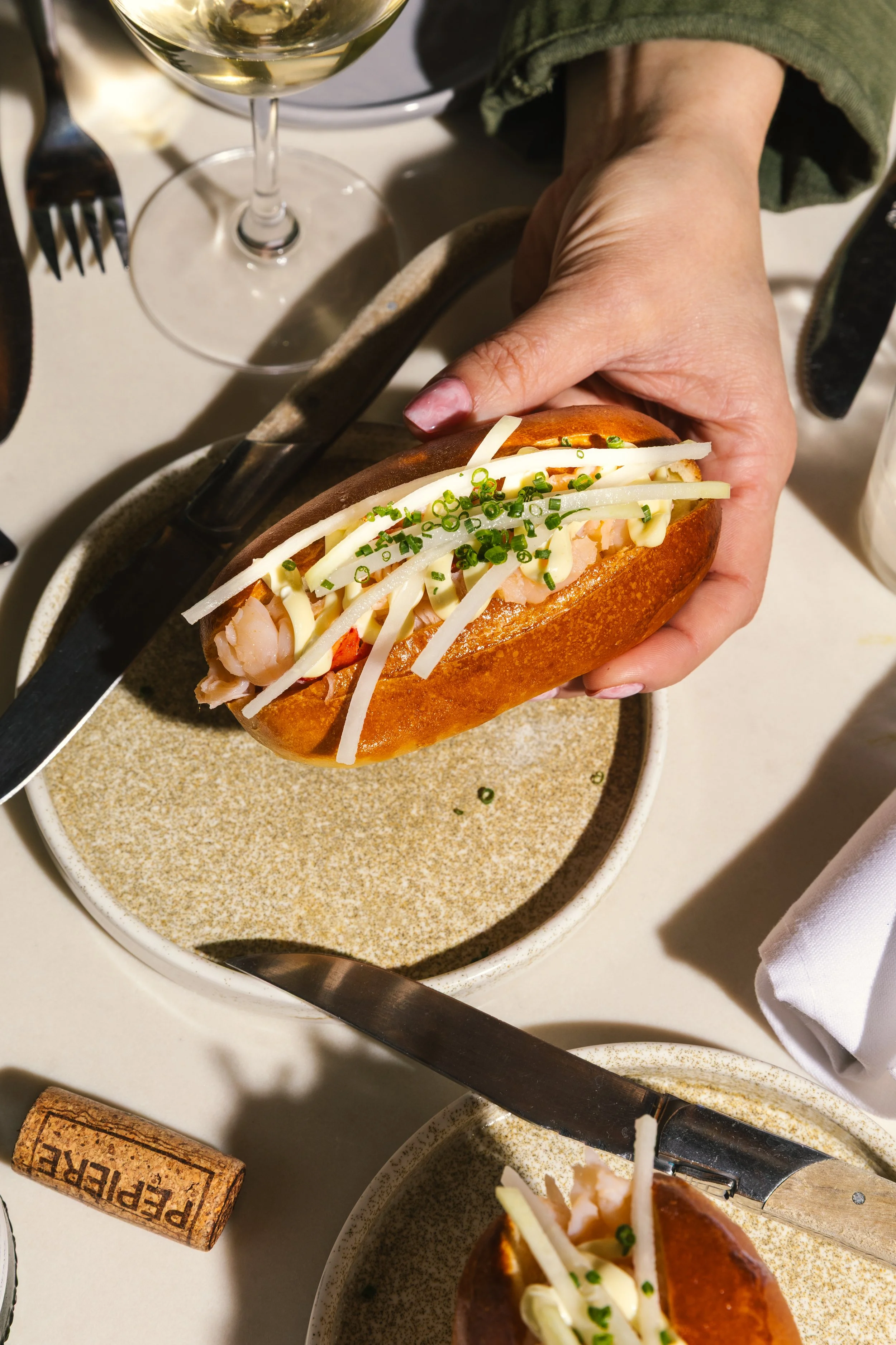 Close-up of a person holding a sandwich filled with shrimp, shredded cheese, and chopped green onions, with a glass of white wine nearby.