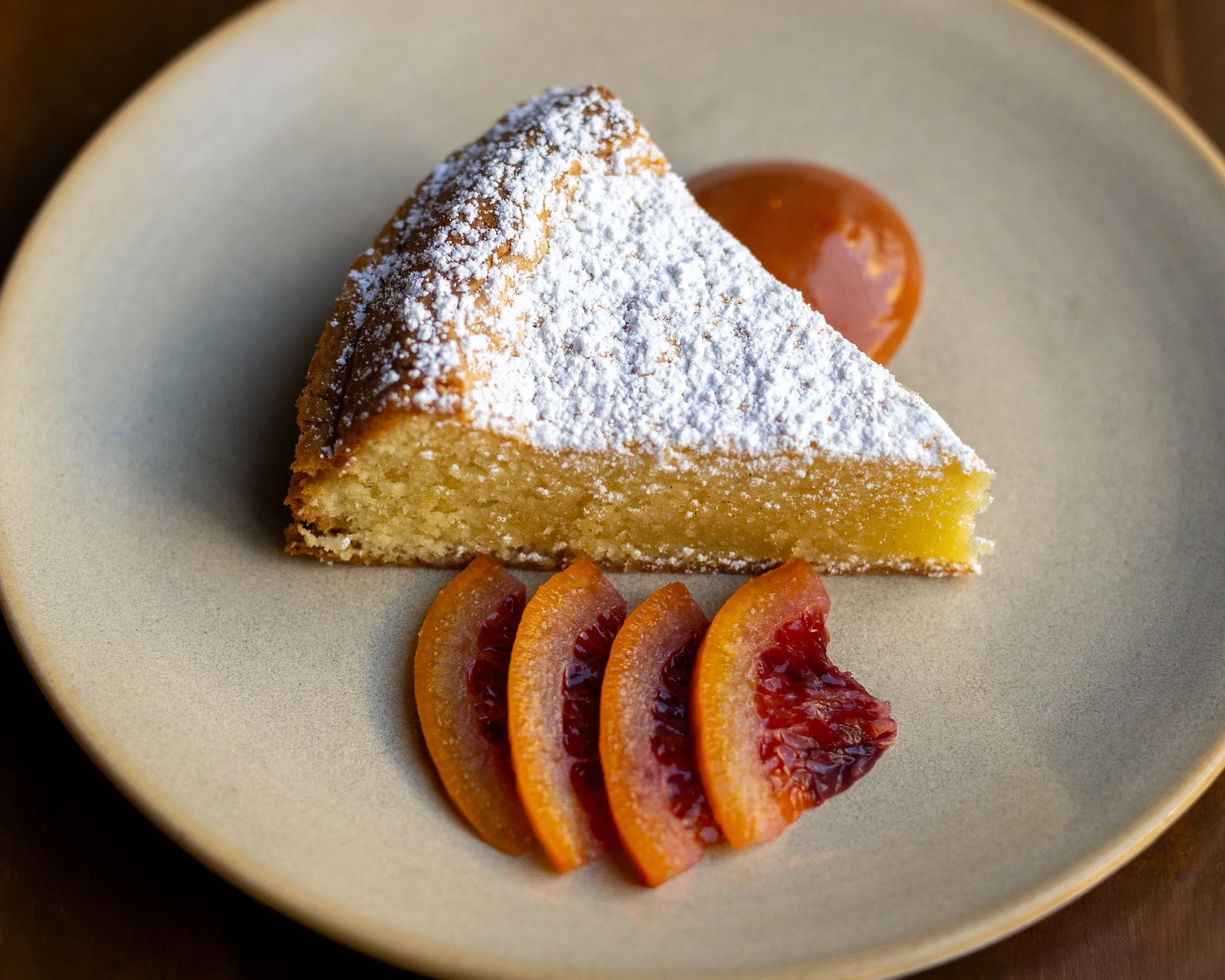A slice of lemon cake dusted with powdered sugar, garnished with blood orange slices and a dollop of caramel sauce on a beige plate.