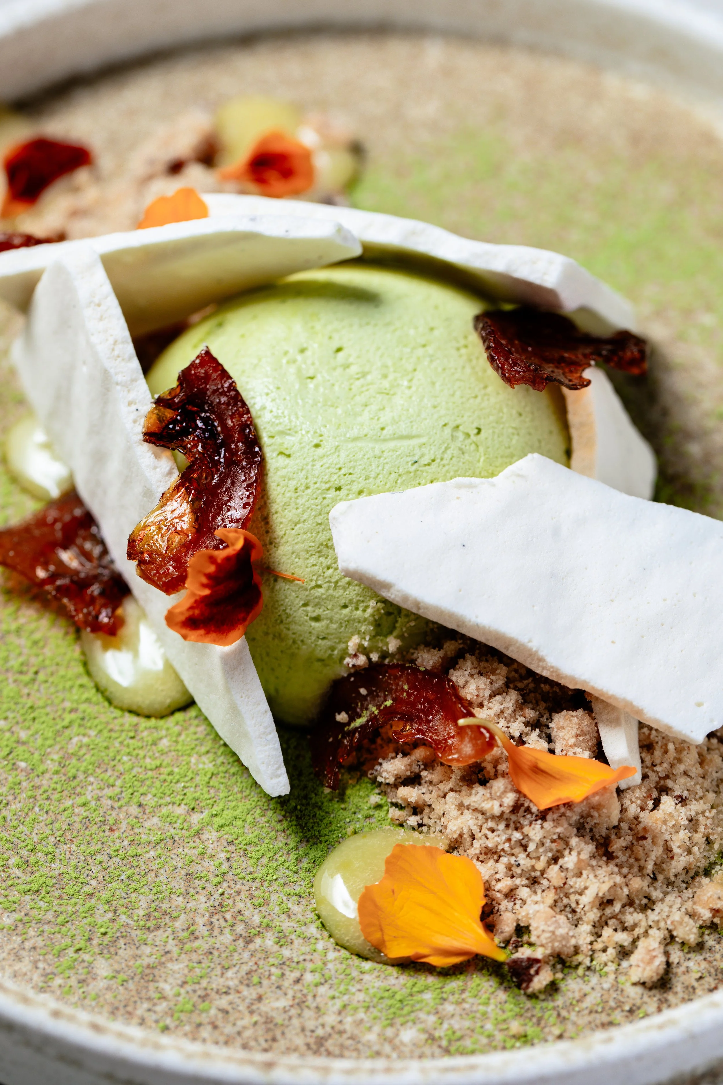 Deconstructed dessert with green ice cream, crispy white meringue, bacon strips, edible flower petals, and crumbled biscuit on a textured green surface.