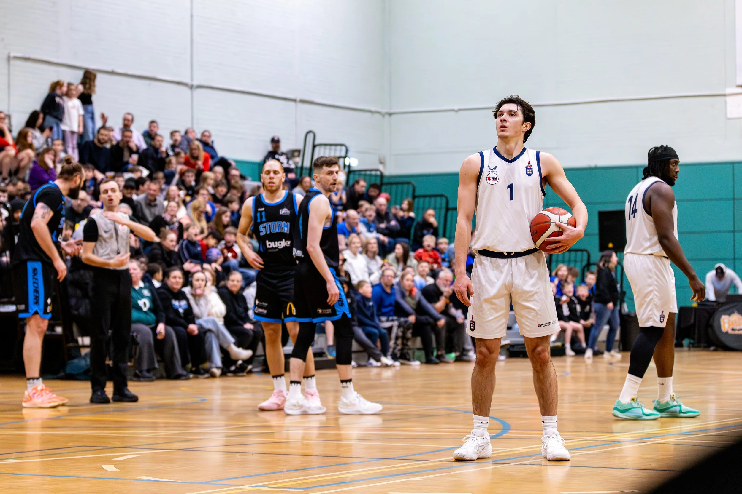 Basketball players on the court with one holding a basketball, and spectators in the background.
